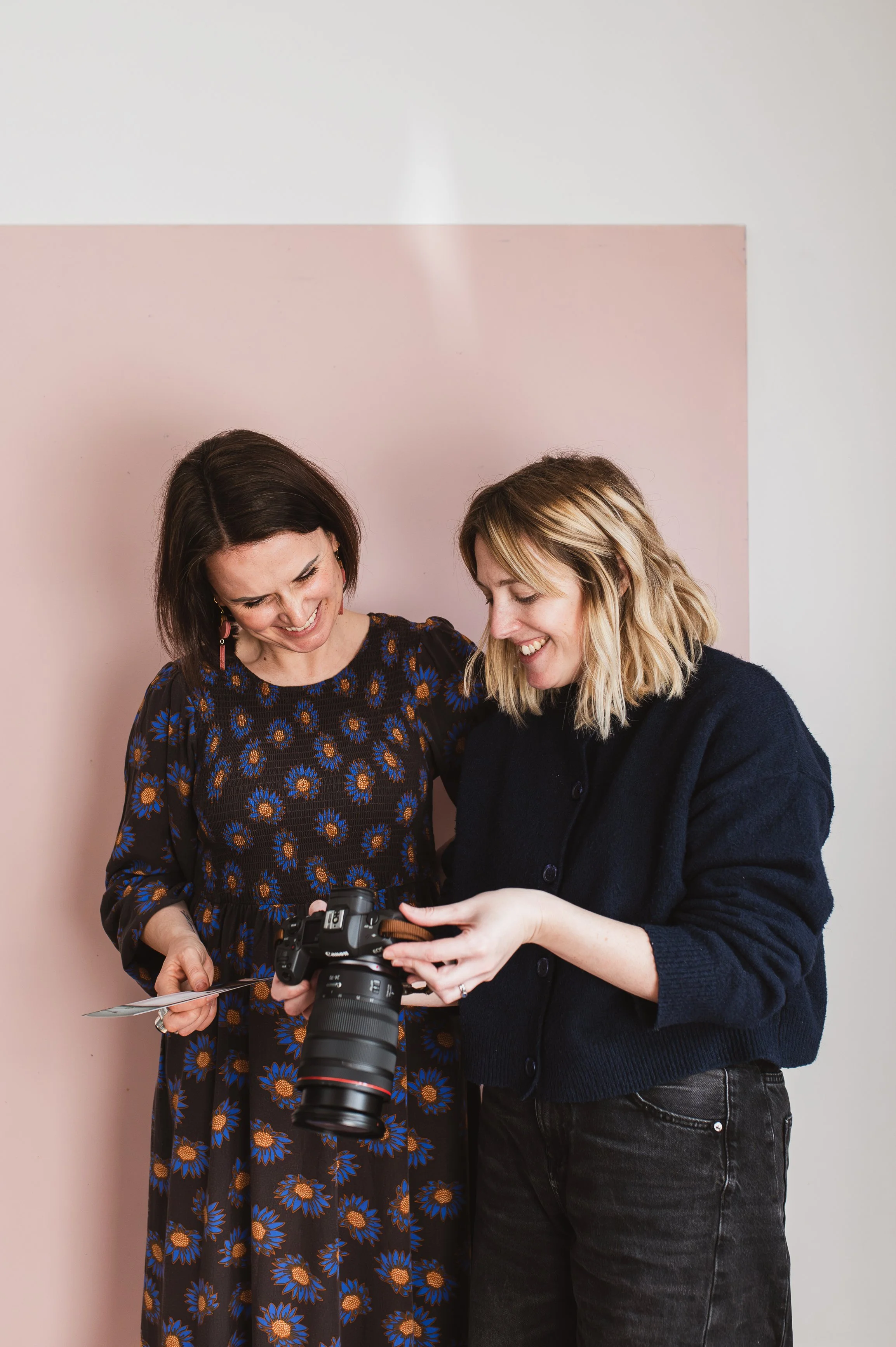  Lady with blonde hair wearing a dark blue jumper and black trousers, standing next another lady in a dress. They are looking at photos on a camera 