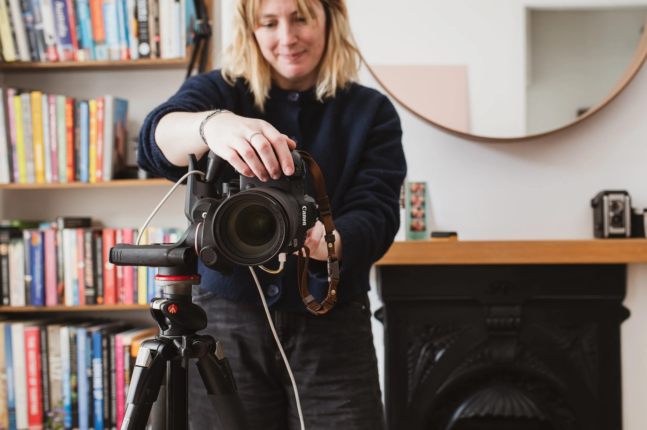 Lady with blonde hair standing behind her camera and taking photos. She is stood in a living room setting with books in the backround and a mirror and fireplace to the right.