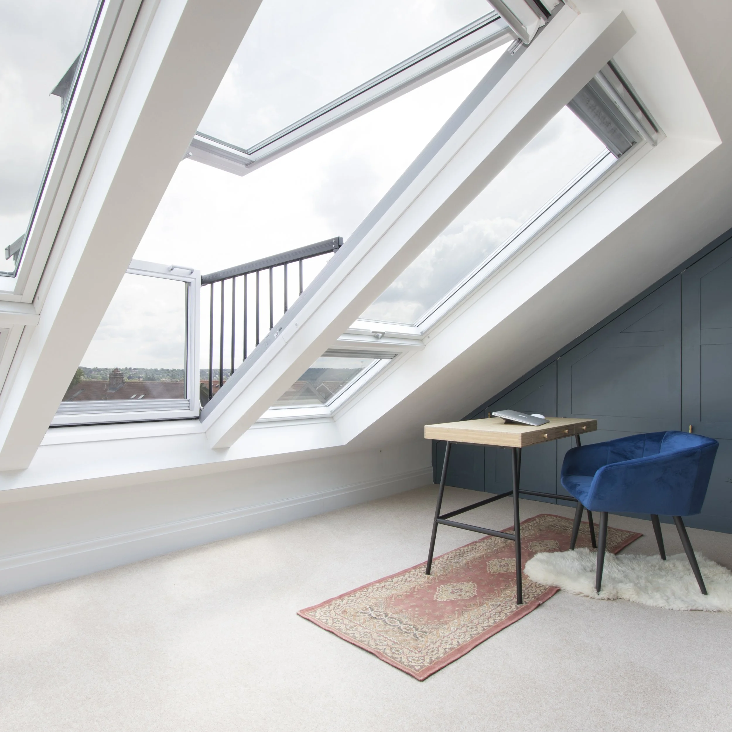 Attic room with sloped ceilings and large tilted skylight windows showing a cloudy sky and rooftops outside. The room has light beige carpet, a small wooden desk with black metal legs, a blue velvet armchair. Bristol Interior Photographer Faye Hedges