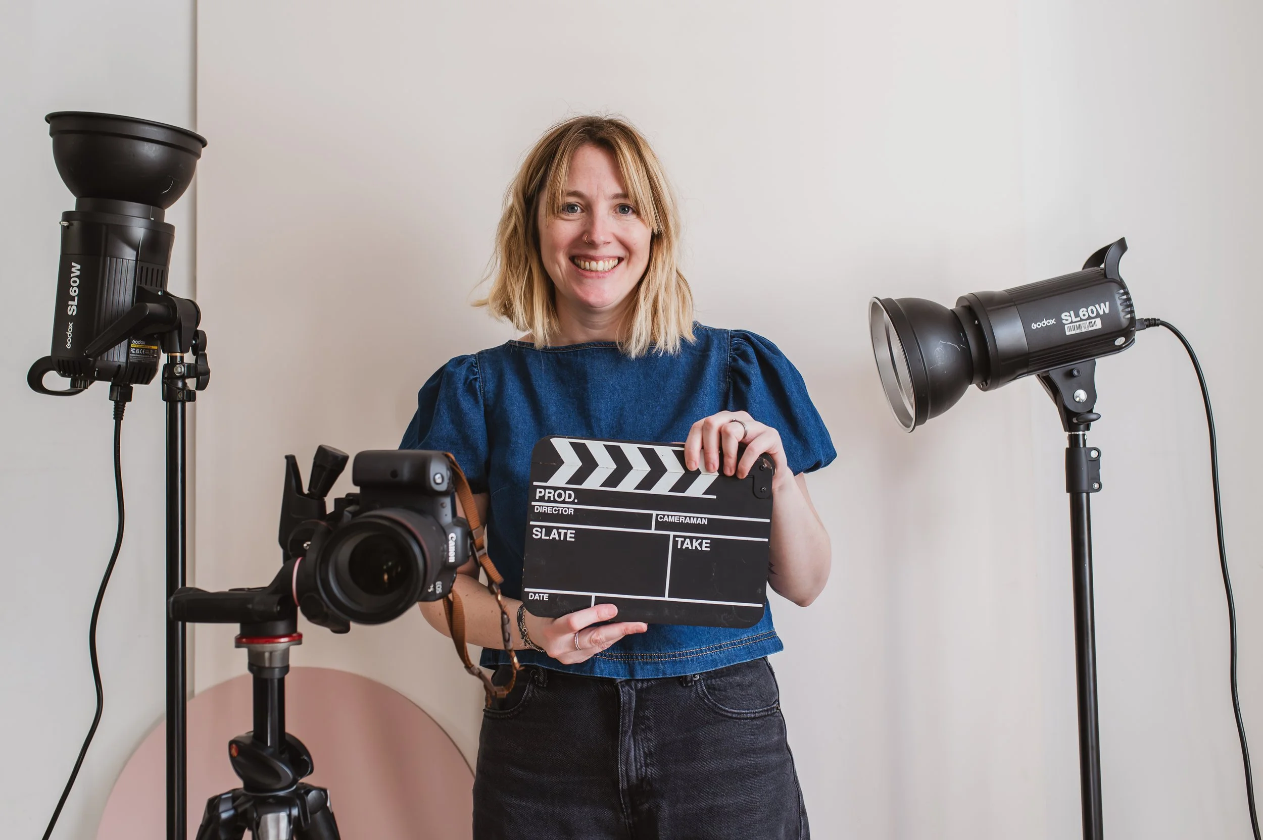 Lady with blonde hair smiling at the camera. She is wearing a blue top and holding a clapperboard. There is camera equipment all around her.