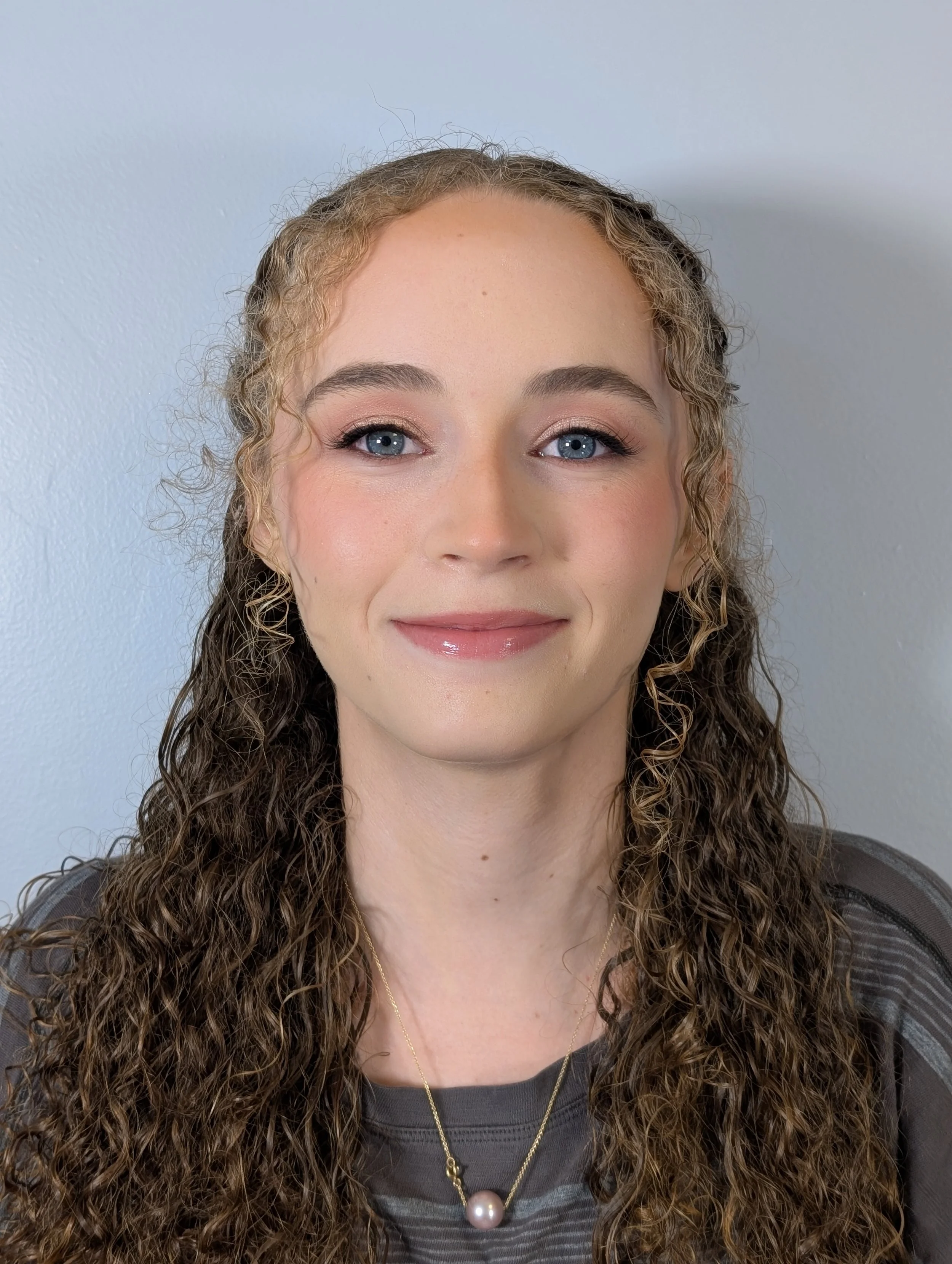 A woman with curly hair, blue eyes, and a light pink lipstick, smiling slightly, wearing a black top, with layered necklaces, standing against a plain white wall.