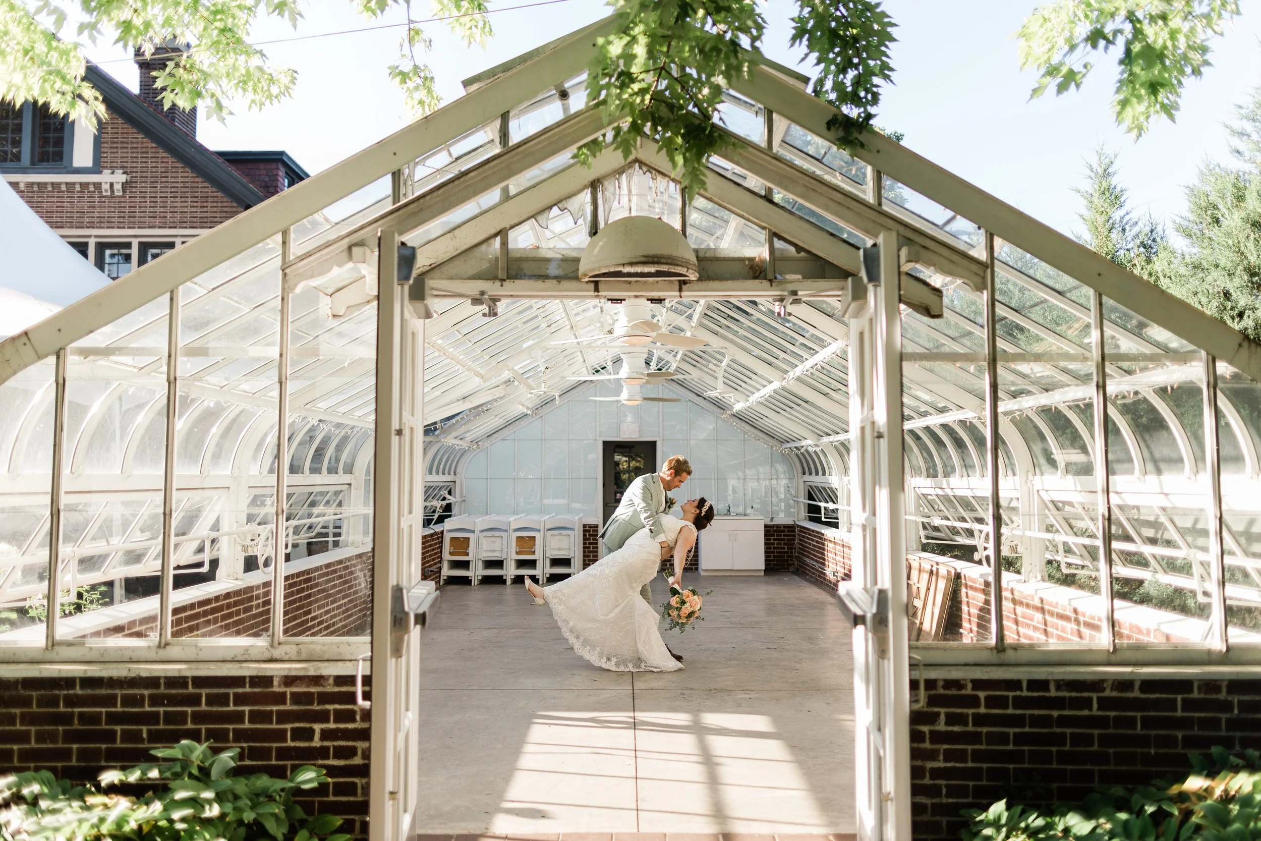 A wedding couple in a greenhouse with the bride being dipped by the groom, surrounded by natural sunlight and greenery.