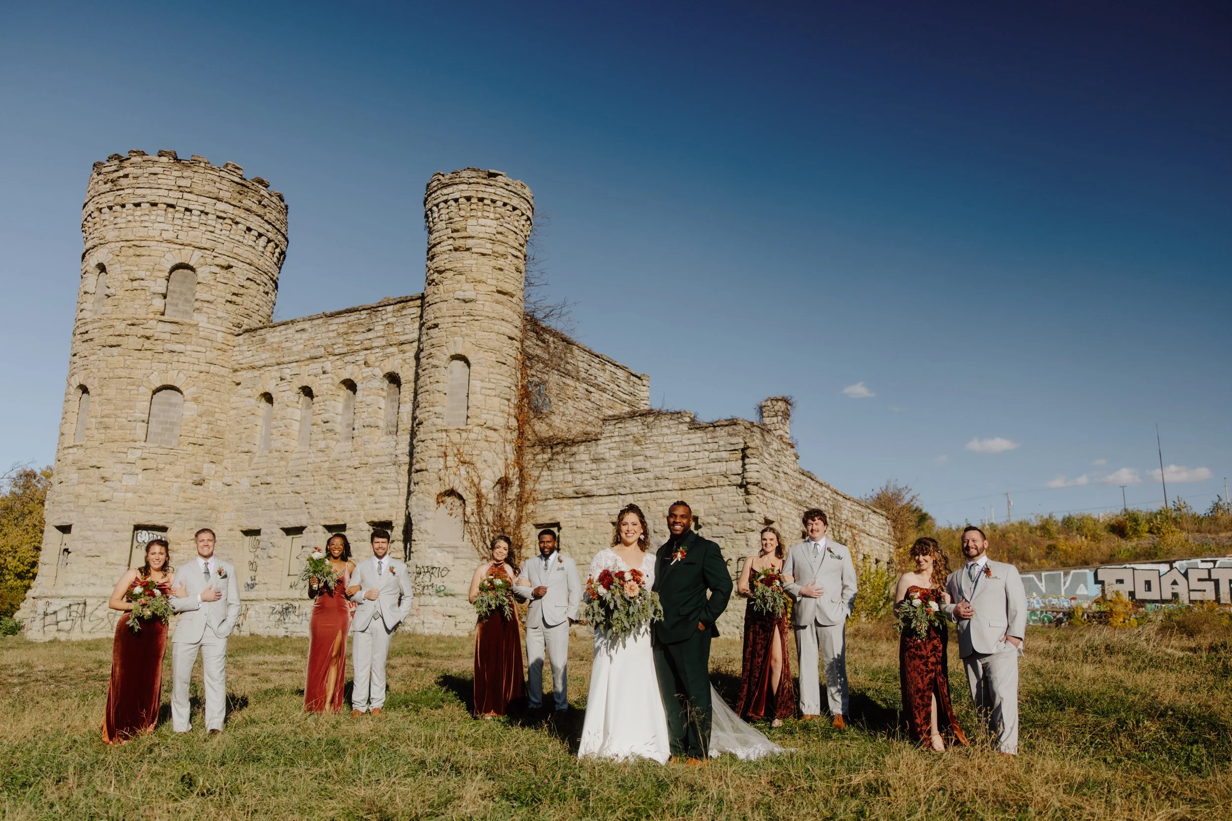 A large group of wedding guests, including the bride and groom, standing outdoors in front of an old stone castle with tall towers under a clear blue sky.