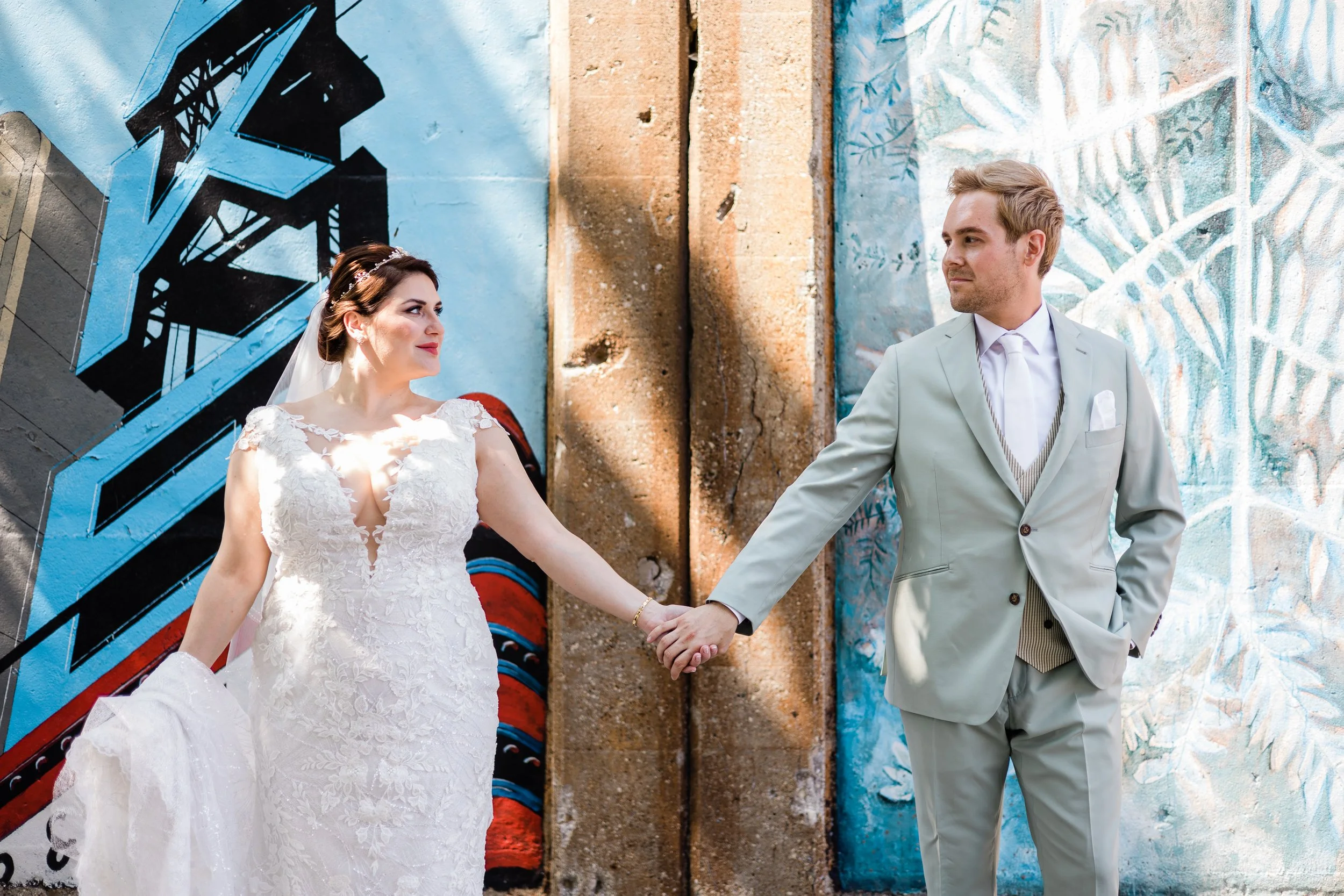 A bride and groom holding hands in front of colorful street art, looking at each other.