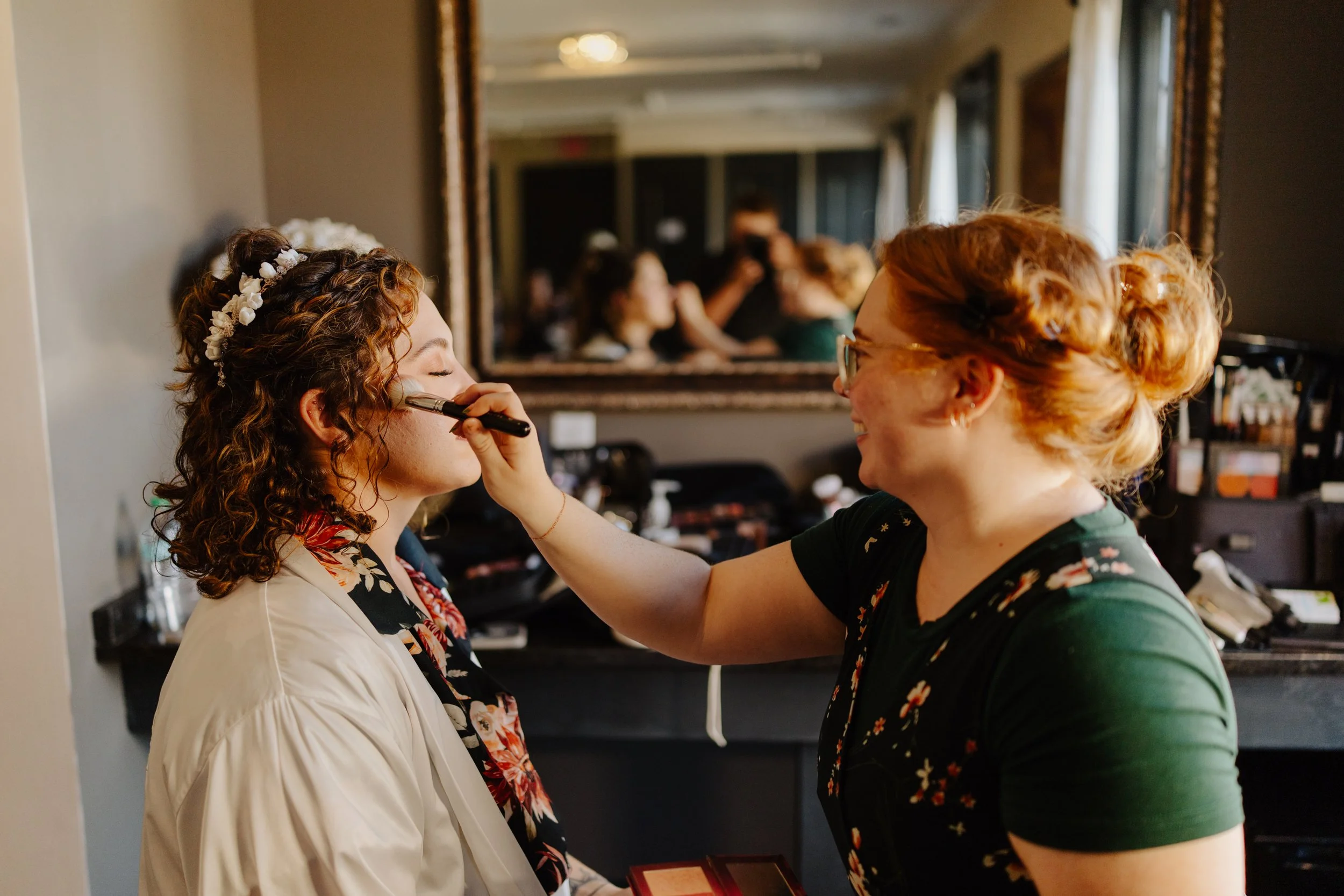 A woman with red hair and glasses applying makeup to another woman with curly brown hair and a flower headband in front of a mirror in a room with makeup products and a person taking a photo in the background.