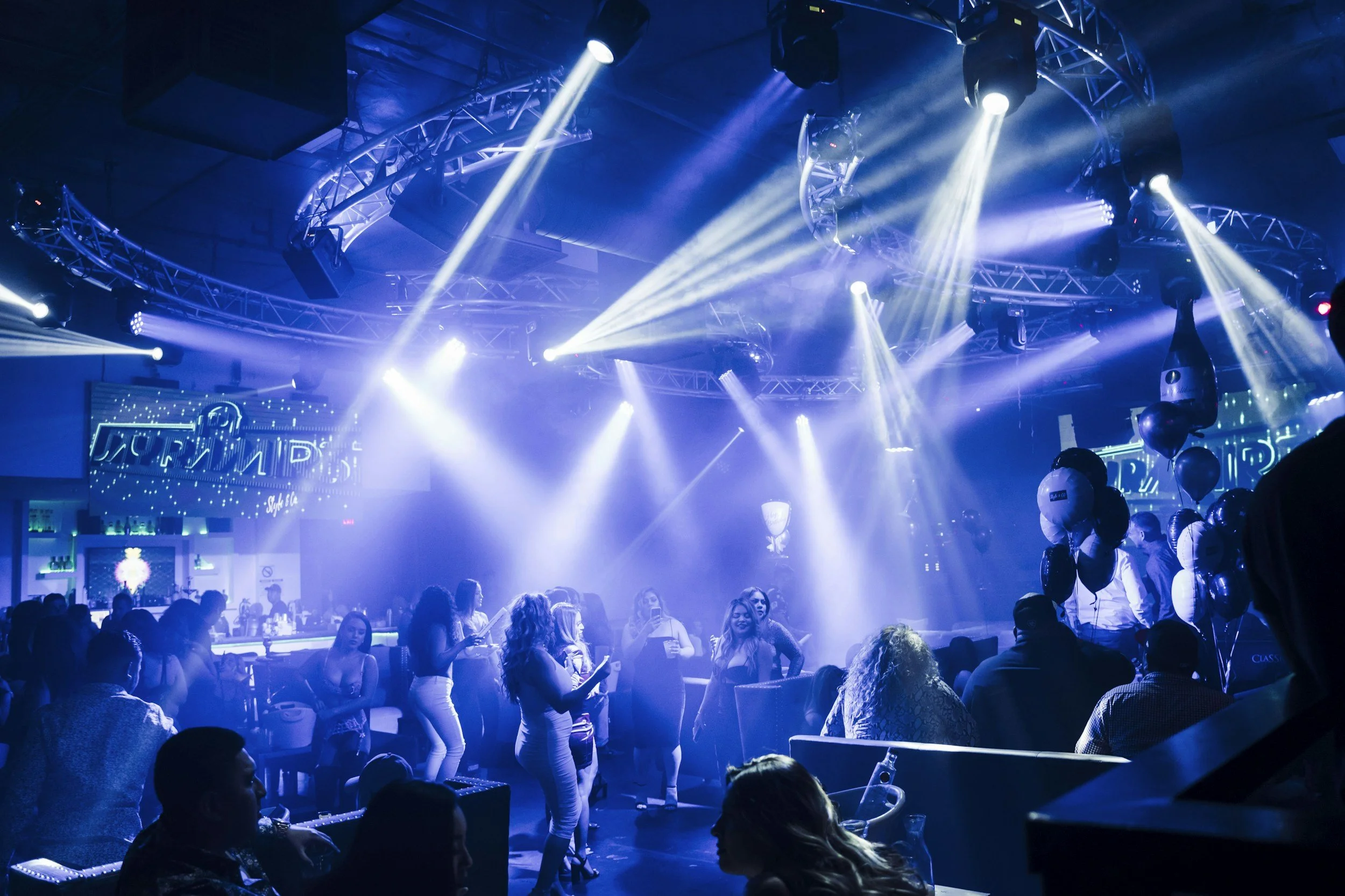 People dancing and socializing in a nightclub with bright blue and white stage lights, a DJ booth, balloons, and digital screens in the background.