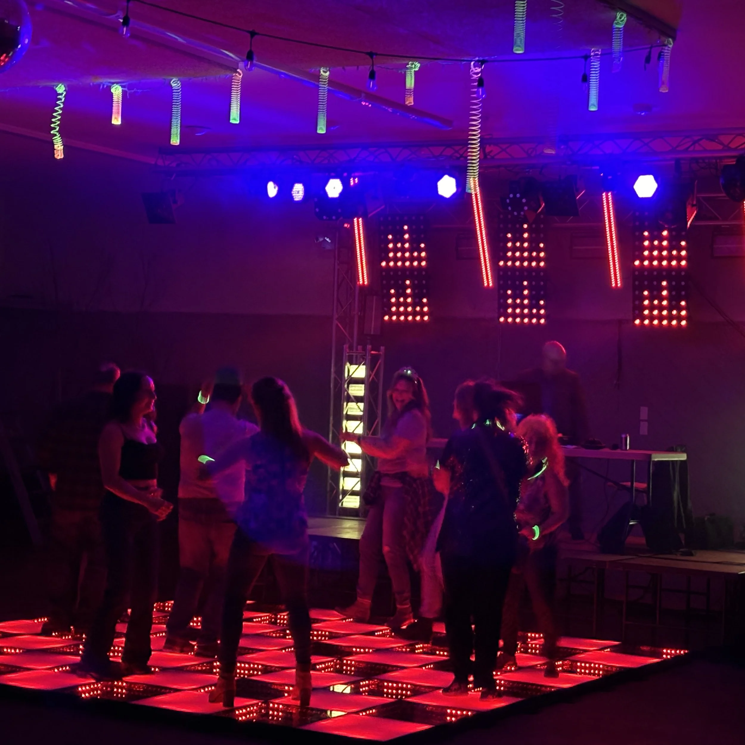 People dancing on an illuminated LED dance floor at a party with colorful stage lighting and a DJ setup in the background.