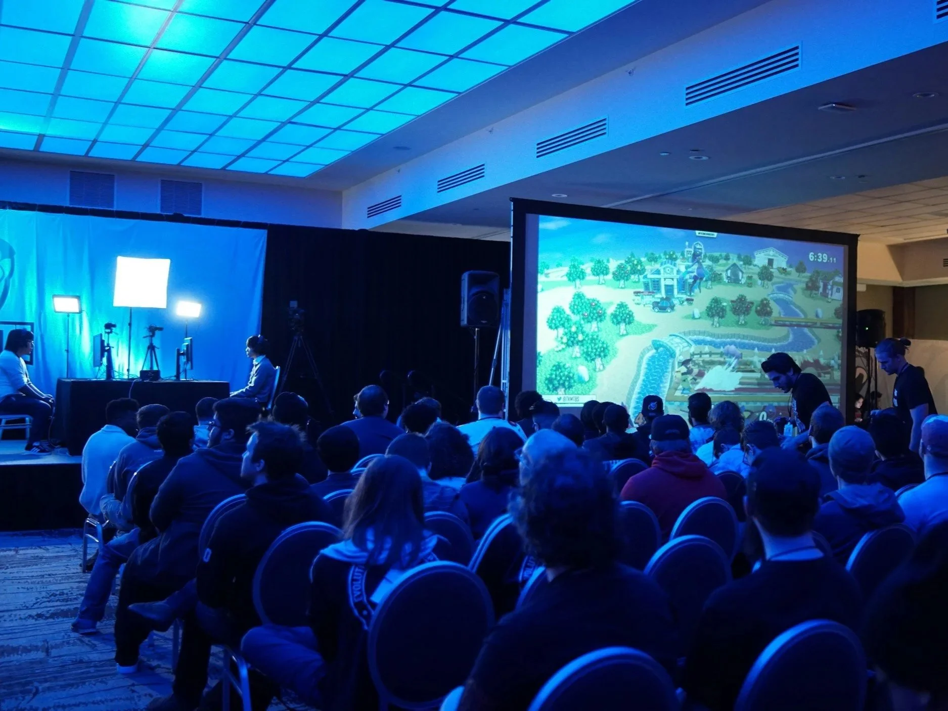 Audience watching a competitive video game tournament on large screen at an indoor event, with players seated at tables and several cameras pointing at the players, illuminated by blue lighting.