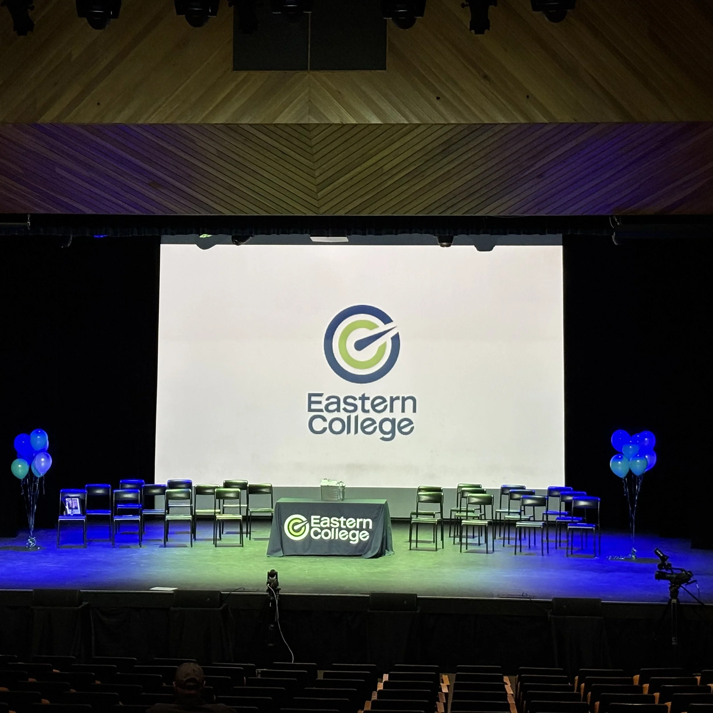 Stage set up for Eastern College, with a large screen displaying the college logo, chairs arranged on stage, balloons on either side, and tables with college branding.