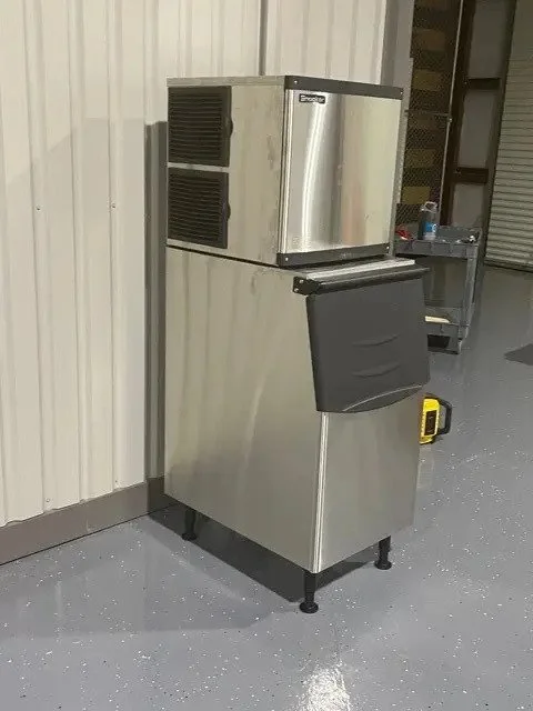 Stainless steel ice machine with a black insulated ice bin underneath, situated next to a white corrugated metal wall in a warehouse or storage room.