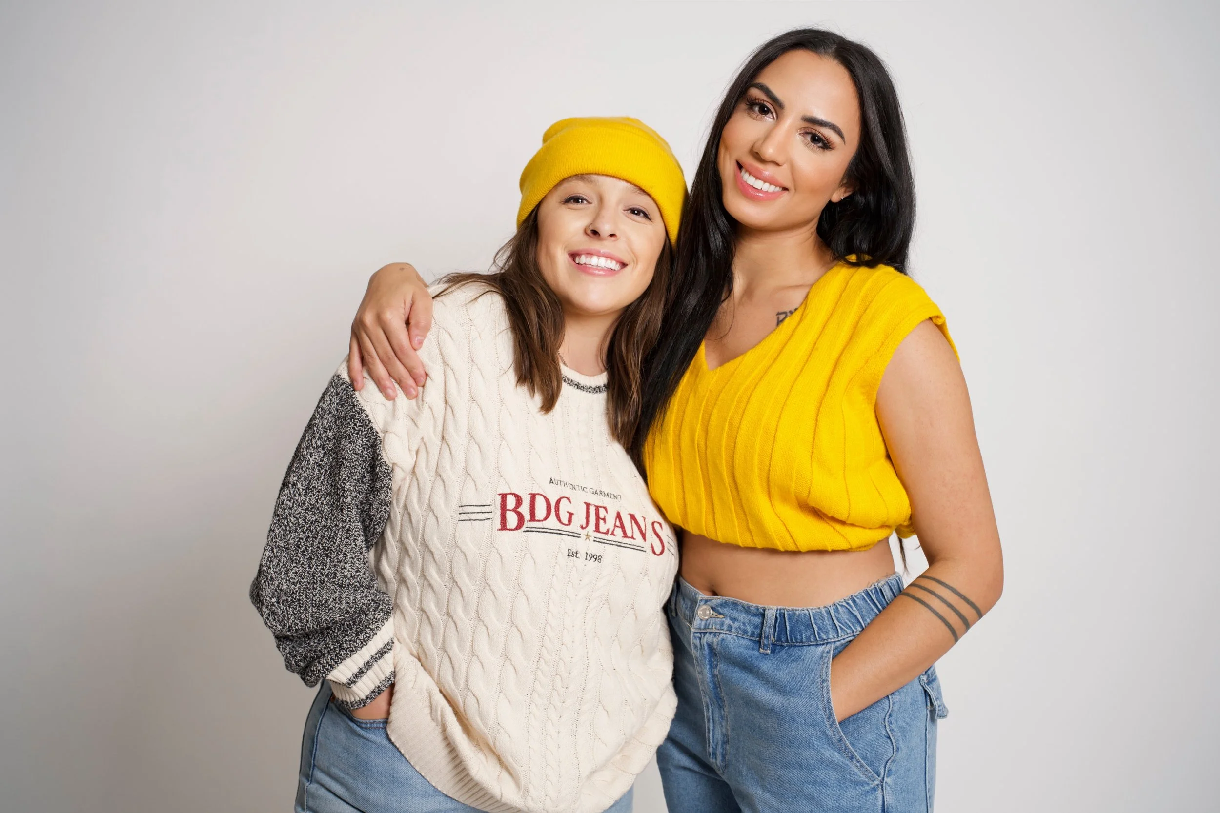 Two women smiling, with one woman wearing a yellow beanie and a white sweater, and the other woman wearing a yellow sleeveless top and jeans, standing close together against a plain white background.