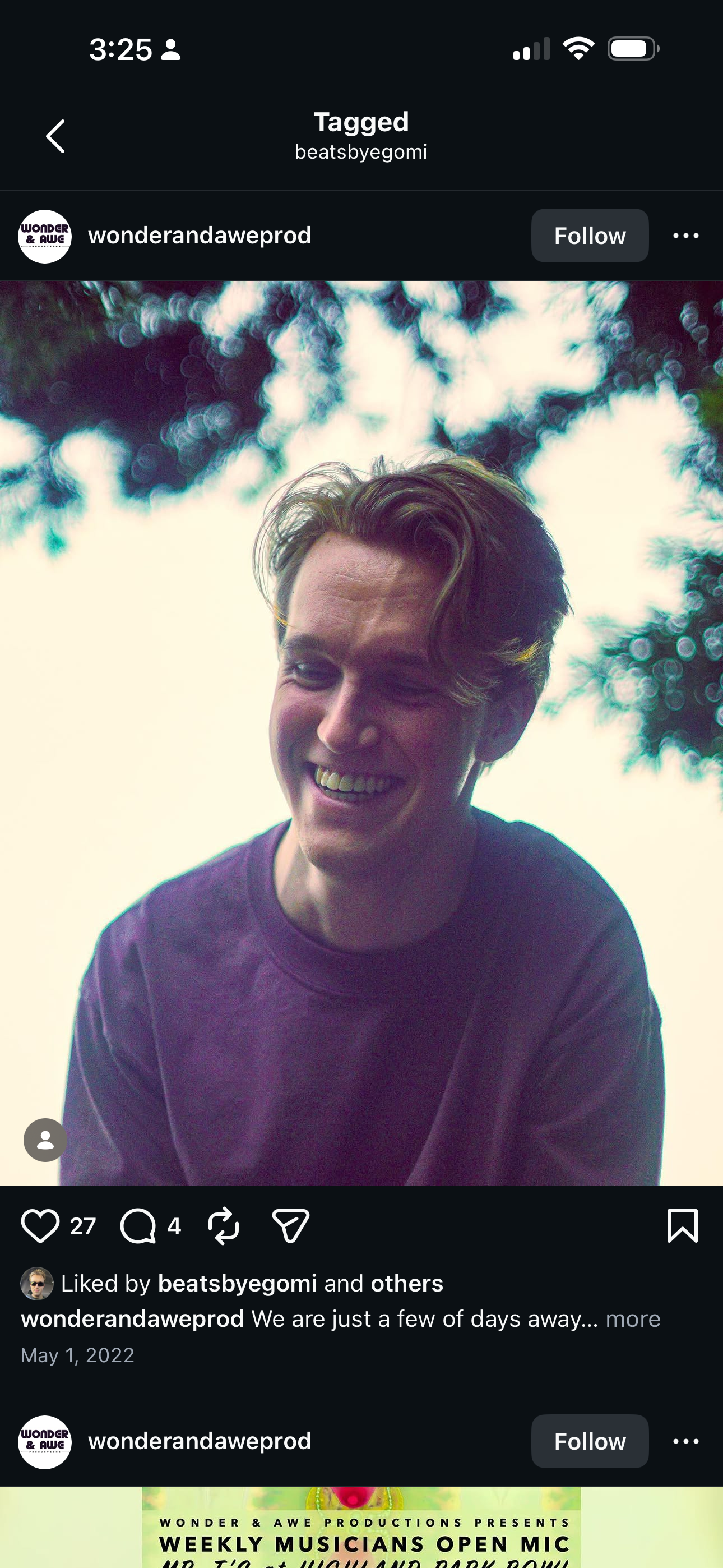 A young man with wavy hair smiling outdoors against a bright sky with blurred tree leaves in the background.