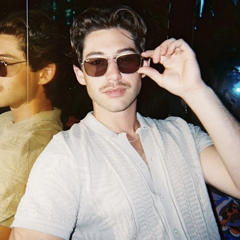 A young man wearing sunglasses, a light-colored button-up shirt, and a chain necklace, posing indoors with a dark background.