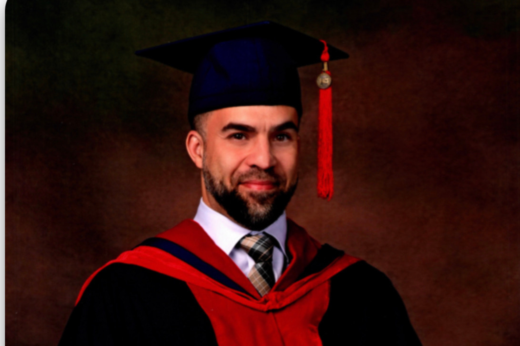 Man in graduation cap and gown, smiling, with a brown background.