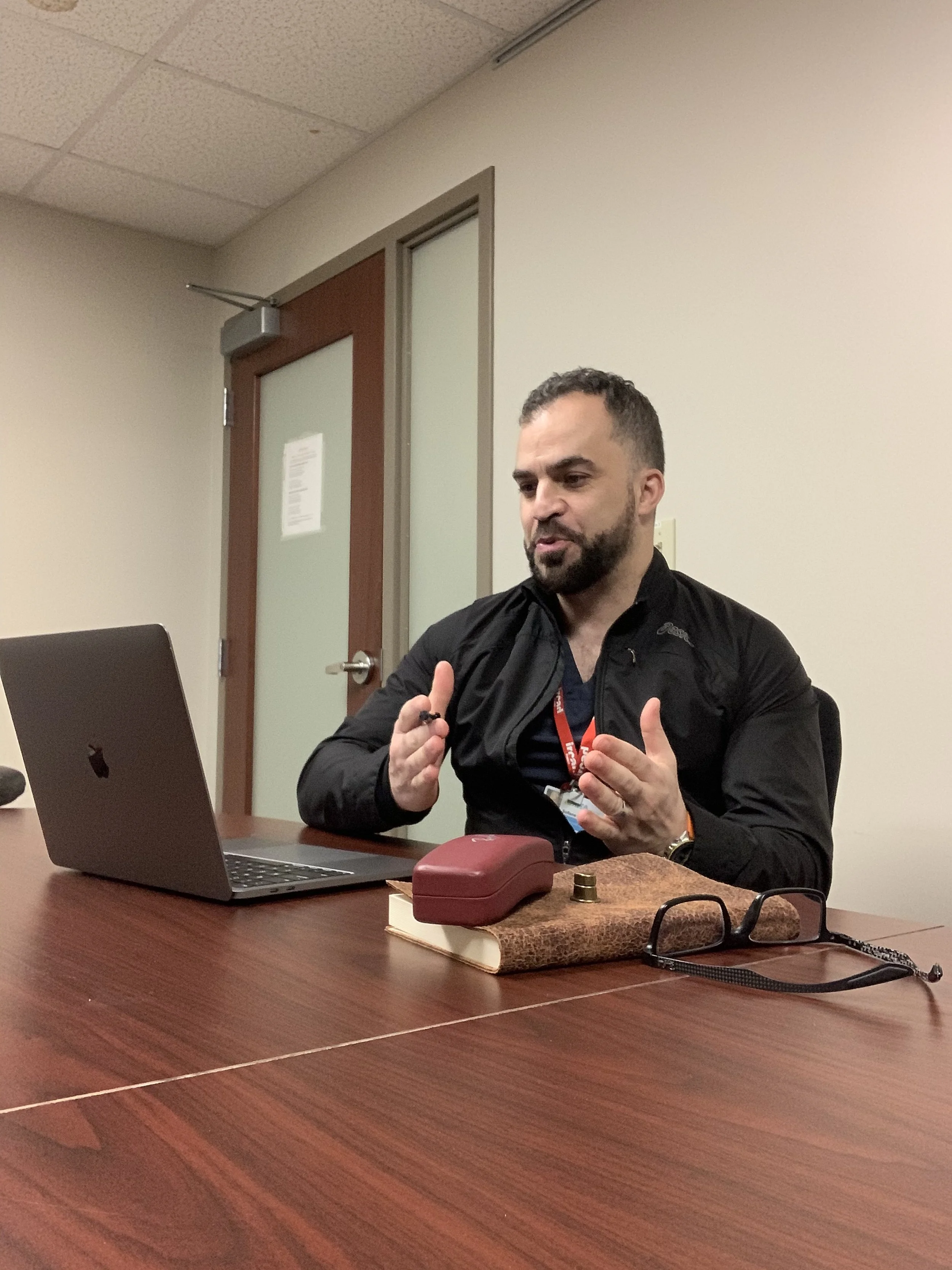 Man with beard and short hair sitting at a table, speaking, with a MacBook, glasses, a book, a small red case, and a camera on the table.