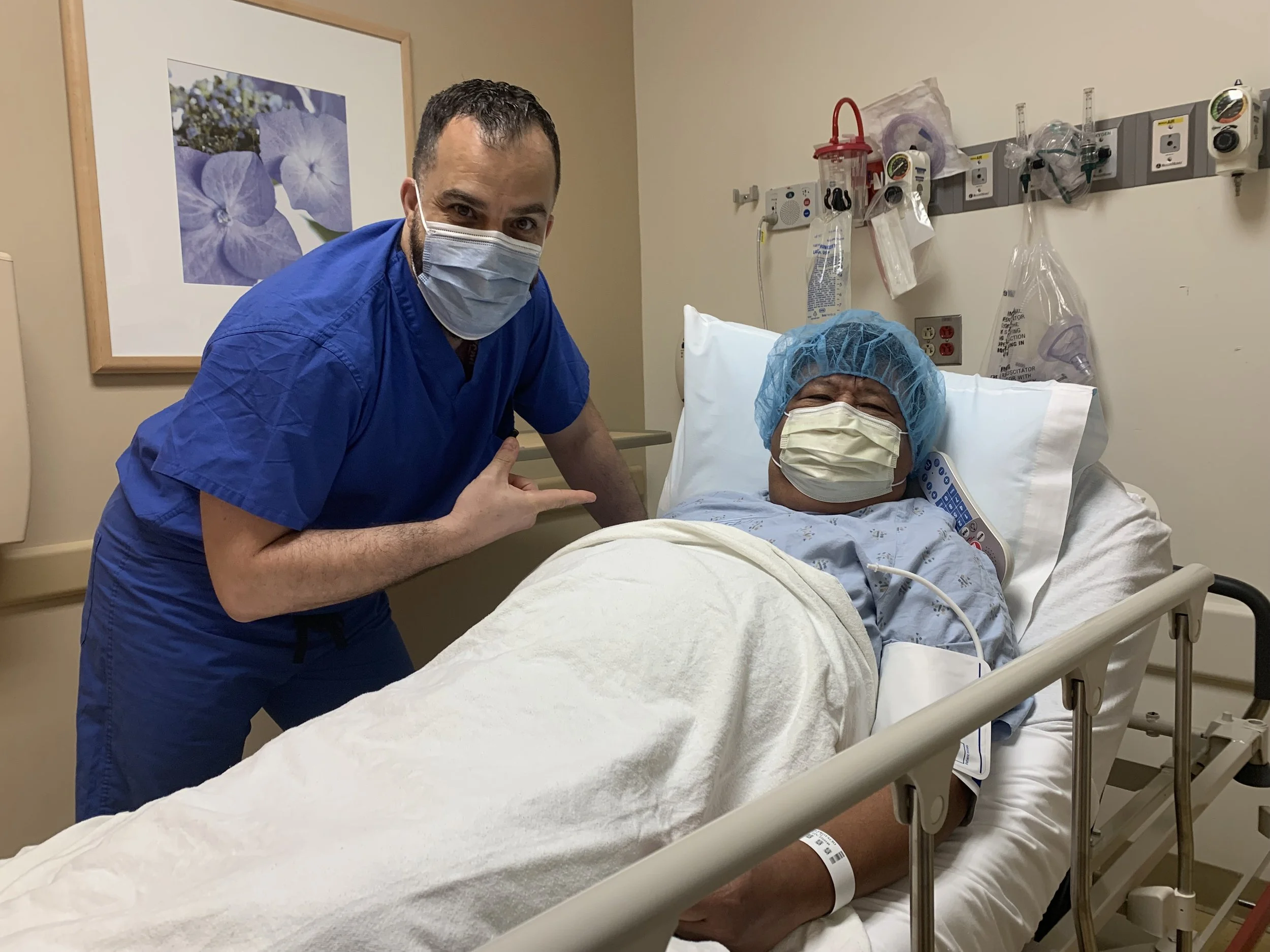 A doctor and a patient in a hospital room. The doctor, wearing blue scrubs and a face mask, is pointing at the patient. The patient, lying in a hospital bed, is wearing a light blue gown, a hair cap, and a face mask. The room has medical equipment and a framed picture of flowers on the wall.