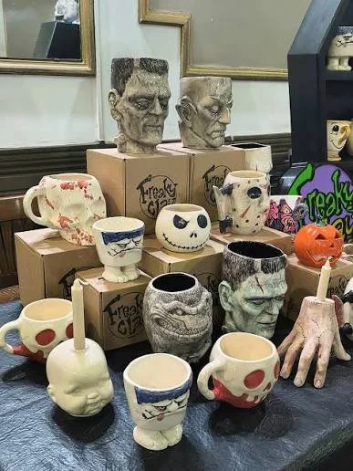 Display of Halloween-themed ceramic mugs and figurines, including skulls, heads, a pumpkin, and a hand, arranged on a table with boxes and mirrors in the background.