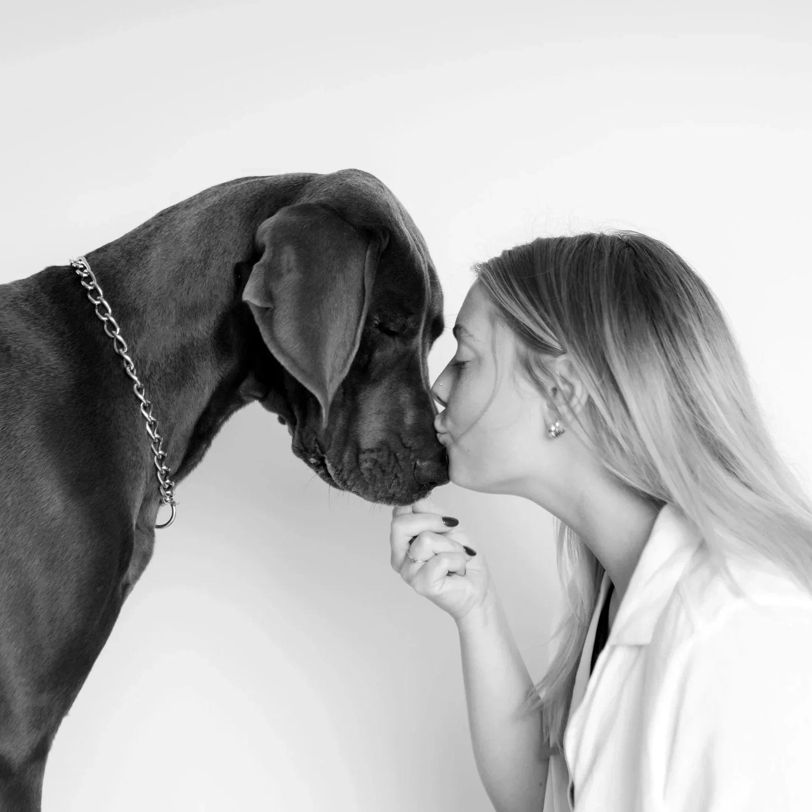 A woman kissing the nose of a Great Dane puppy, each with eyes closed, in a black and white photo.