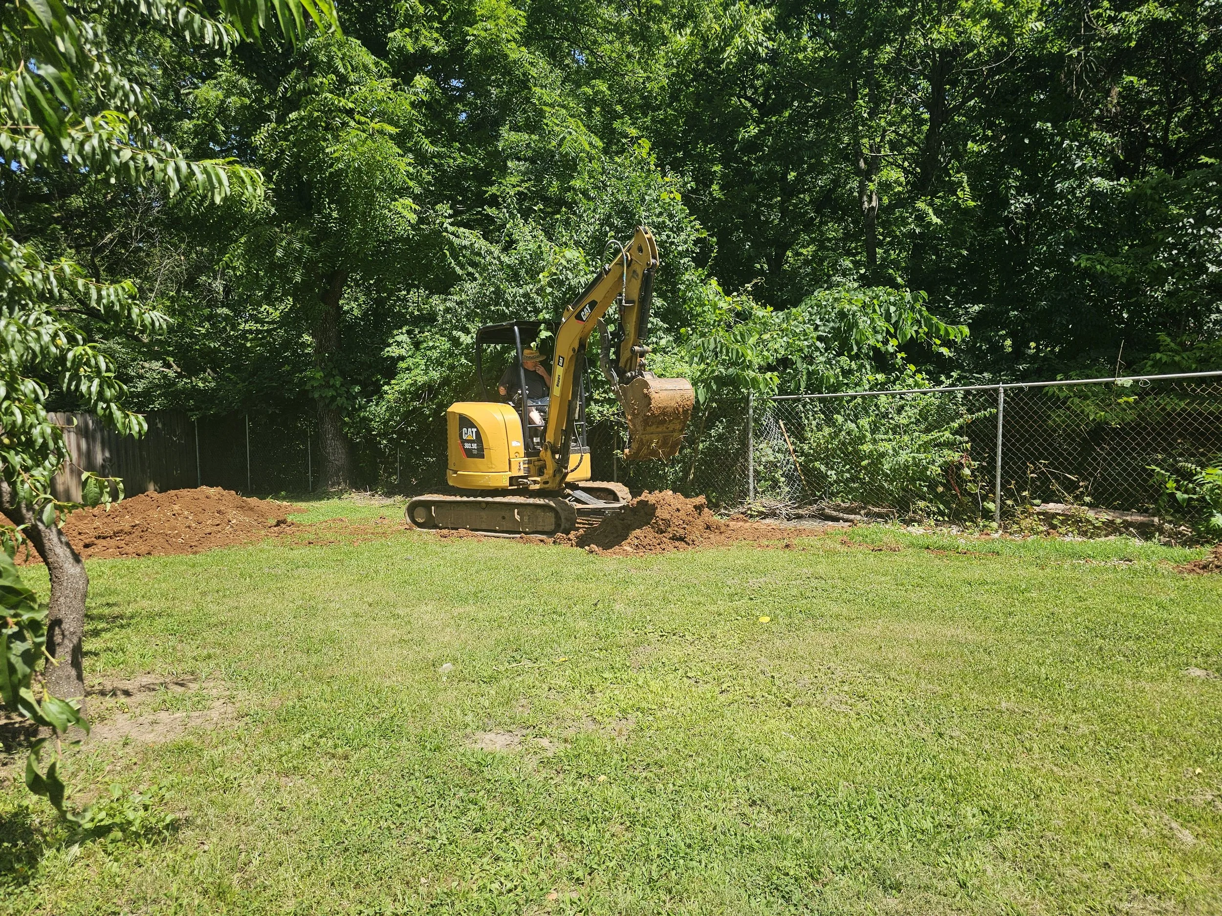 A yellow CAT mini excavator working on digging a hole in a backyard with green grass, trees, and a chain-link fence.