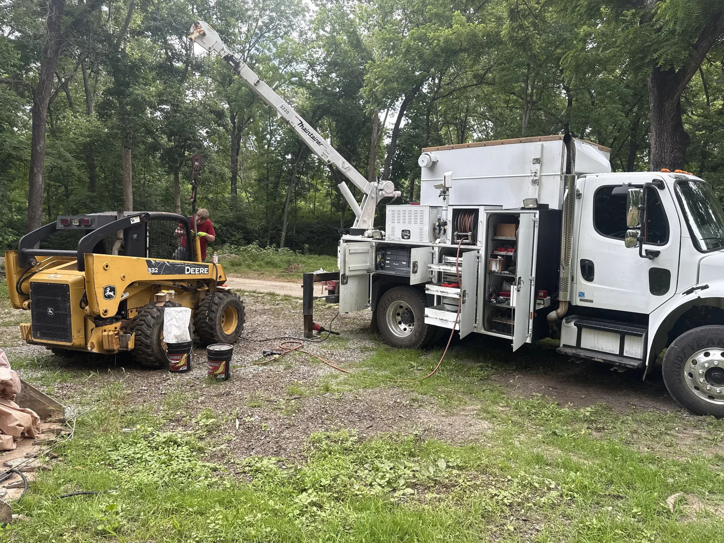 A person in red shirt working with equipment near a large white service truck with open compartments, a crane attached, and a small yellow tractor in a wooded area.
