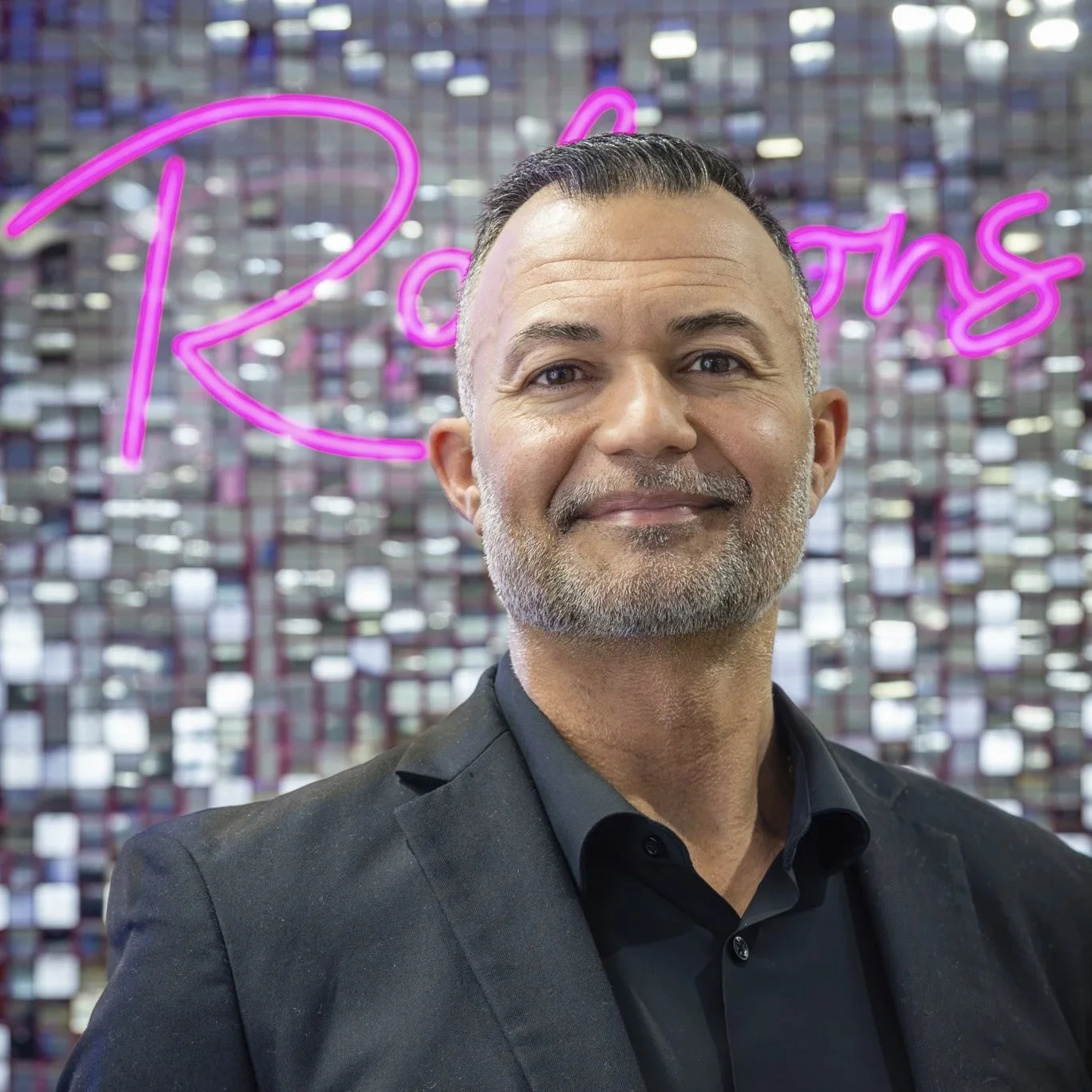 A man with short gray hair and a beard, wearing a black suit and black shirt, standing in front of a reflective mosaic wall with pink neon lights that spell out 'Robson's'.
