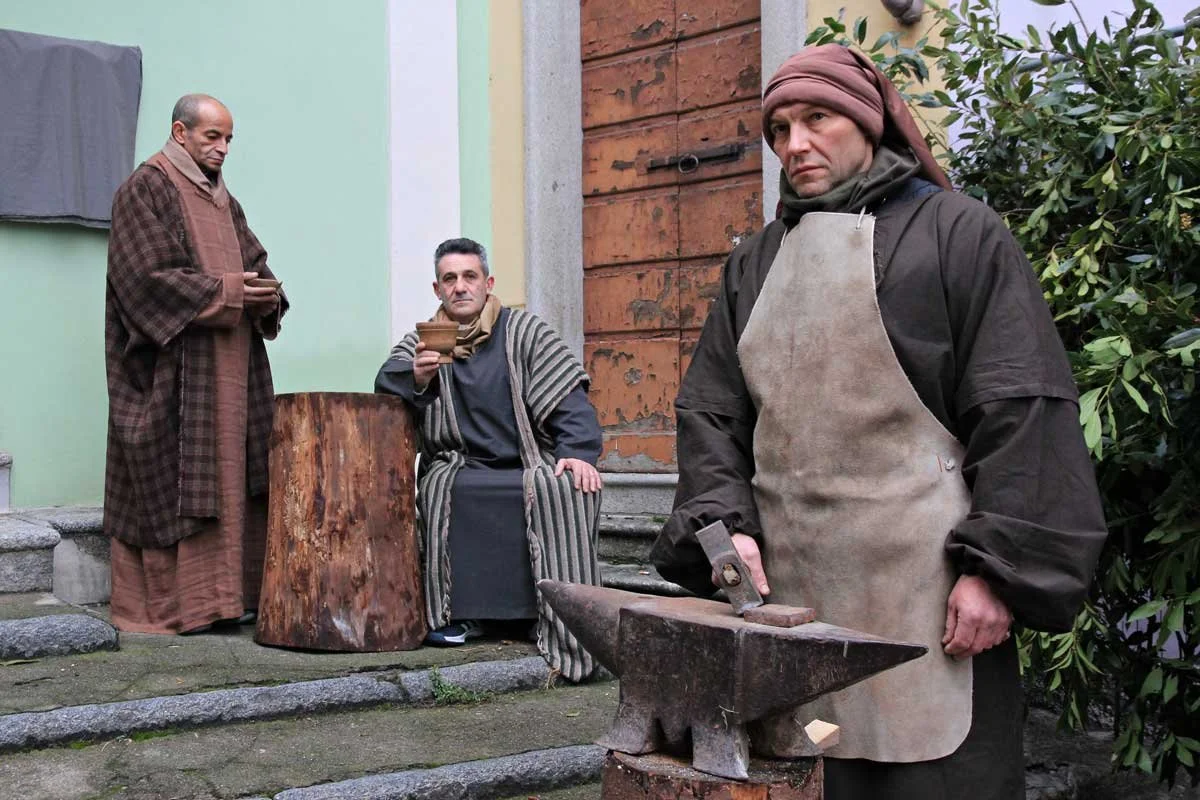 Tre uomini in costumi storici, uno con un martello su un'incudine, un altro che beve da una tazza e un terzo che sta leggendo una pergamena, in un cortile di pietra con un muro di legno.