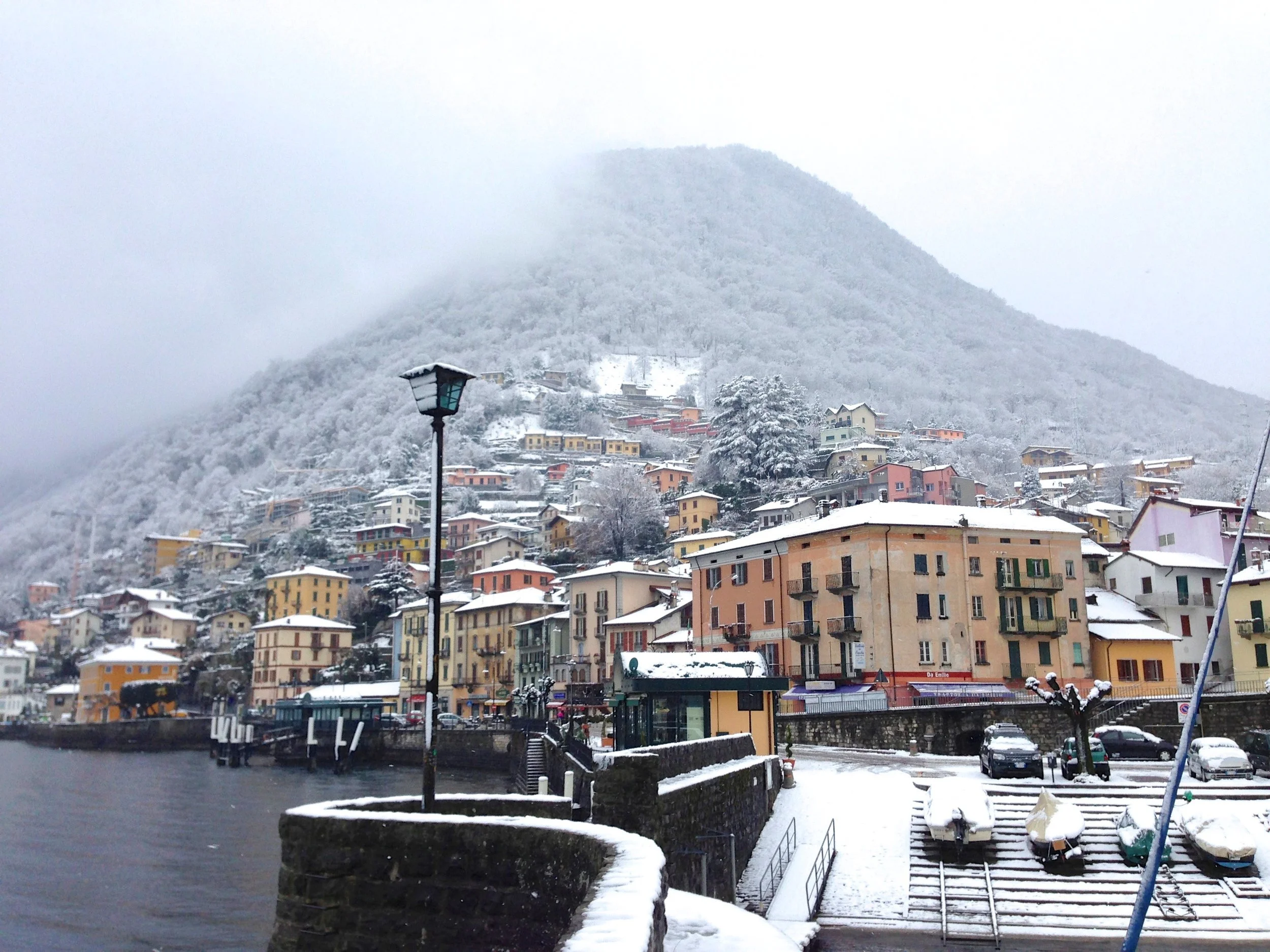 Paesaggio di un villaggio di montagna coperto di neve con case colorate lungo il lago e un monte innevato sullo sfondo.