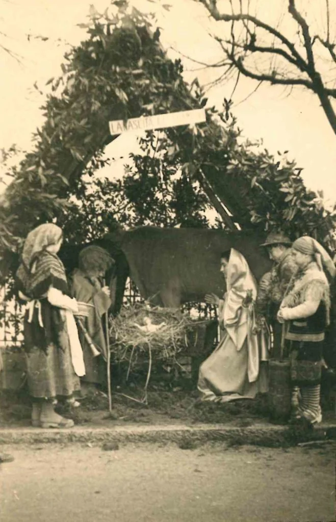 Gruppo di persone vestite con abiti tradizionali, intorno a un cavallo e a un tradizionale presepe, con un arco di foglie e rami sopra. Sullo sfondo un cartello con scritto "LA PASCUA".