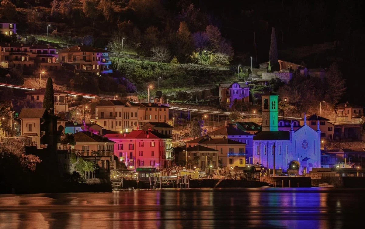 Paesaggio di un villaggio di notte con edifici illuminati, chiesa con torre meridionale colorata, riflessi sull'acqua e colline sullo sfondo