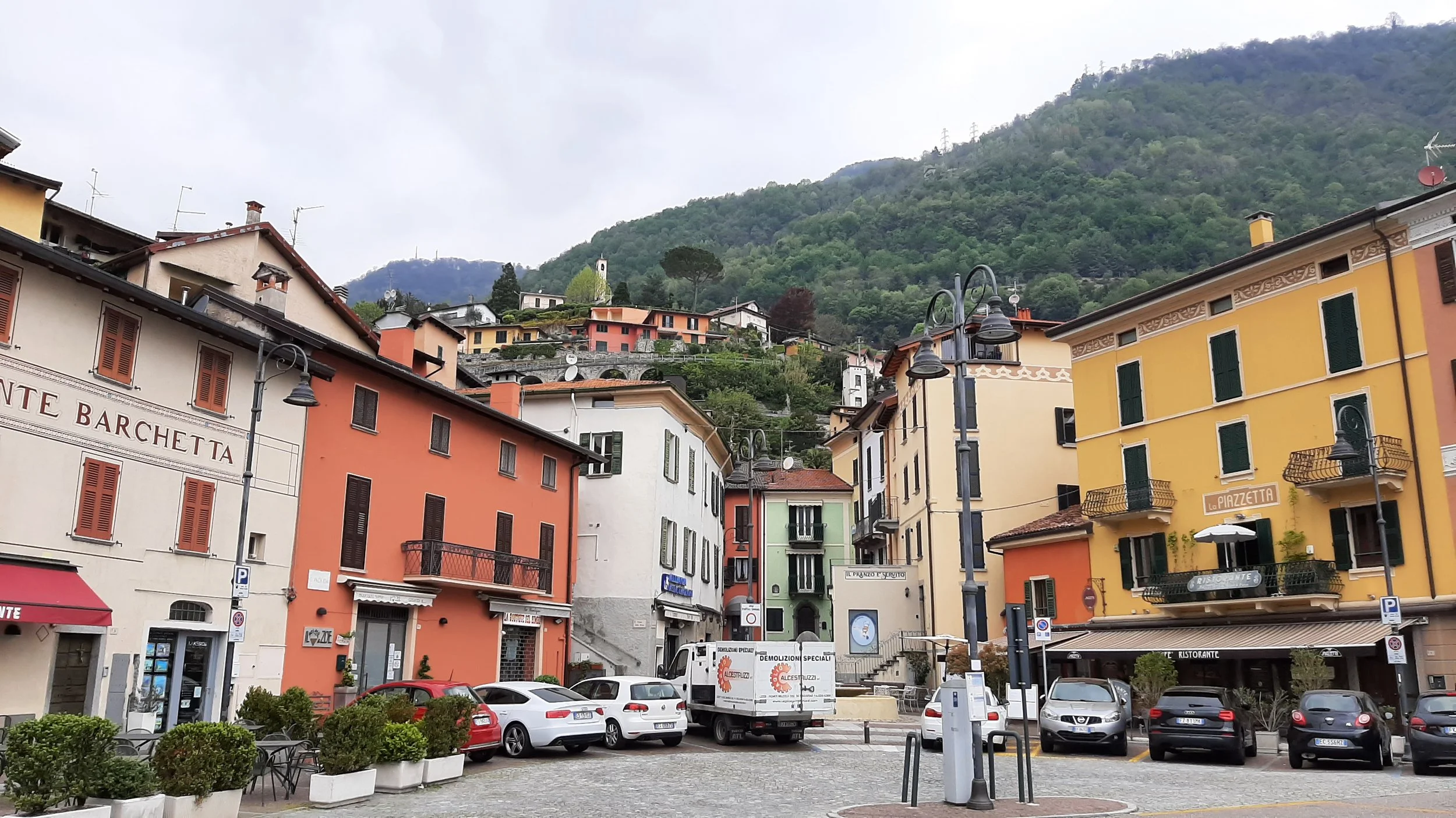 Piazza con edifici colorati e parcheggio, montagne verdeggianti sullo sfondo.