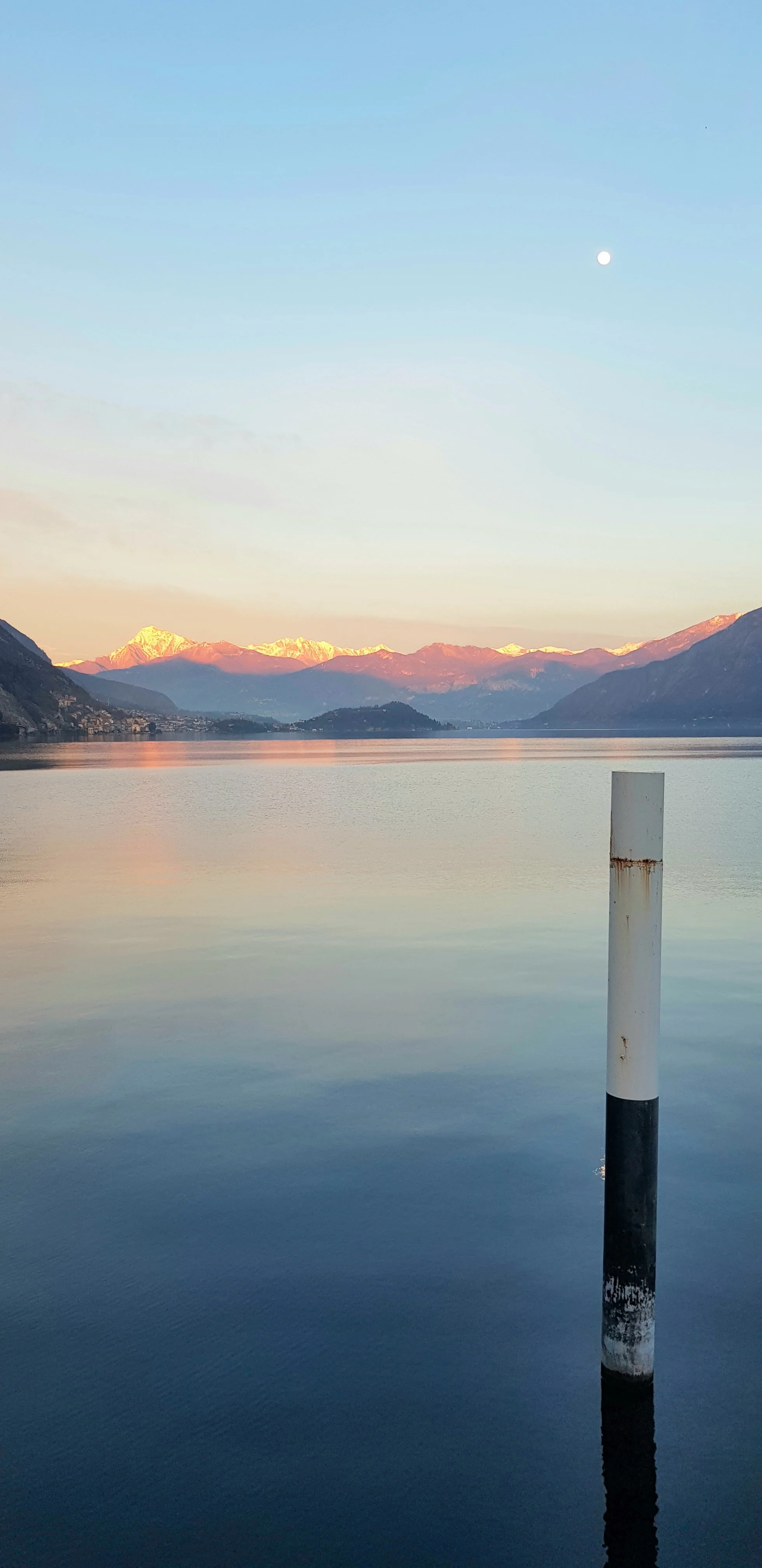 Lago con montagne sullo sfondo e la luna nel cielo, con un palo di segnalazione nell'acqua.