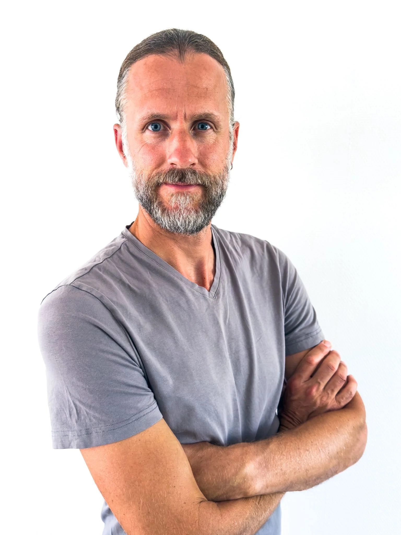 Portrait of a middle-aged man with blue eyes, a beard, and mustache, wearing a gray V-neck T-shirt and standing against a plain white background with arms crossed.