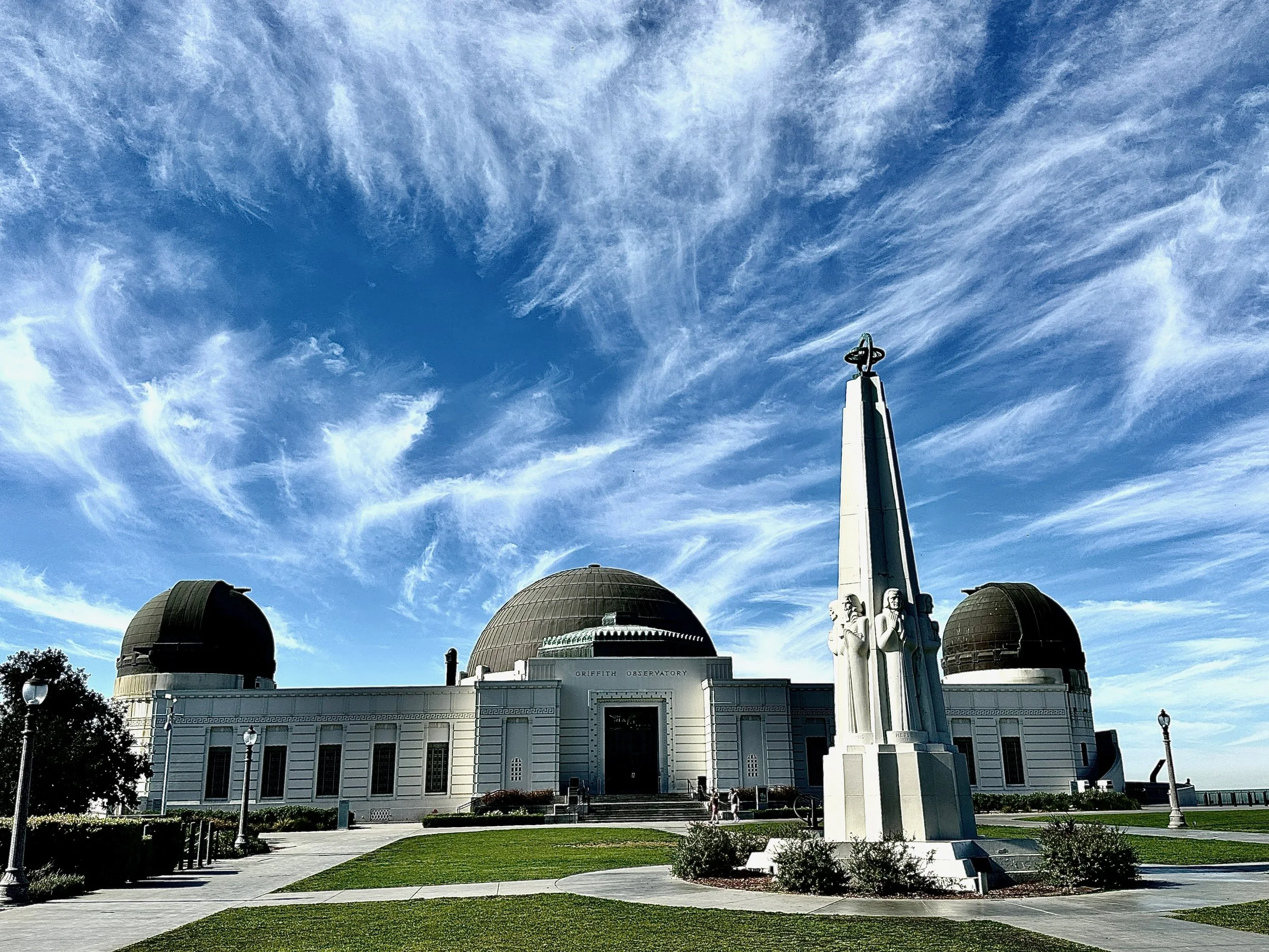 The Griffith Observatory building with its three large domes and a star-shaped monument in front, under a blue sky with wispy clouds.