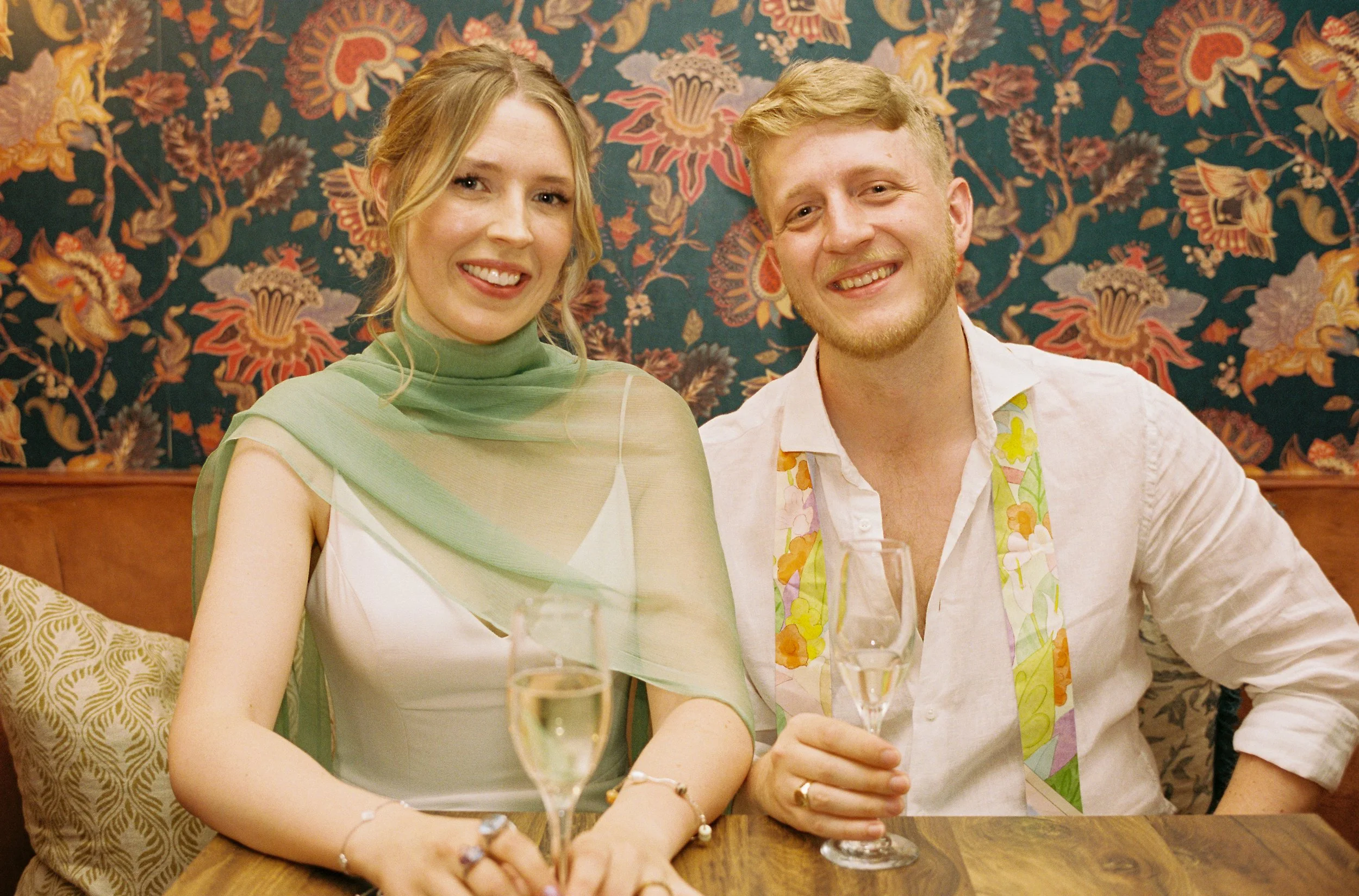 A smiling woman and man sitting at a table with champagne glasses, in a decorated indoor setting with a floral patterned wallpaper.
