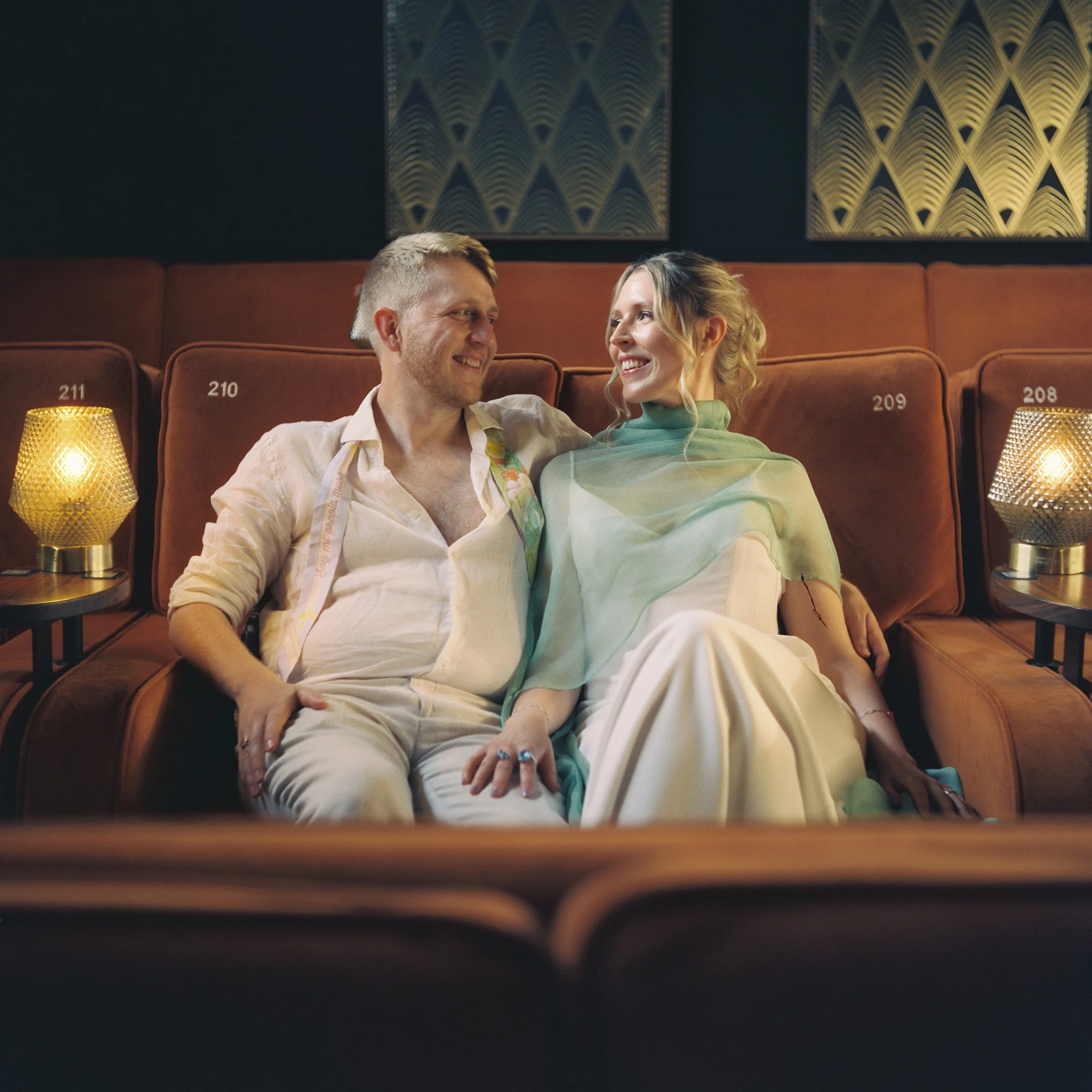 A husband and wife sitting on theater seats, smiling at each other, with warm ambient lighting and decorative wall panels in the background.