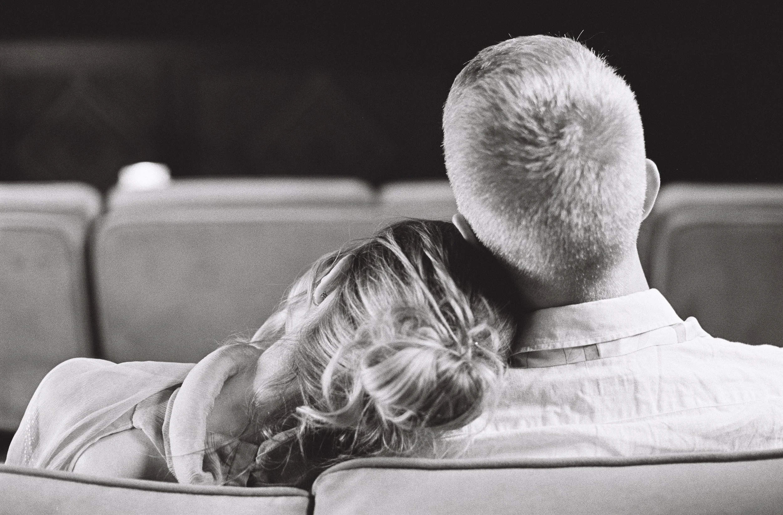 A black and white photo of a man and woman sitting closely in a public space, with the woman resting her head on the man's shoulder.