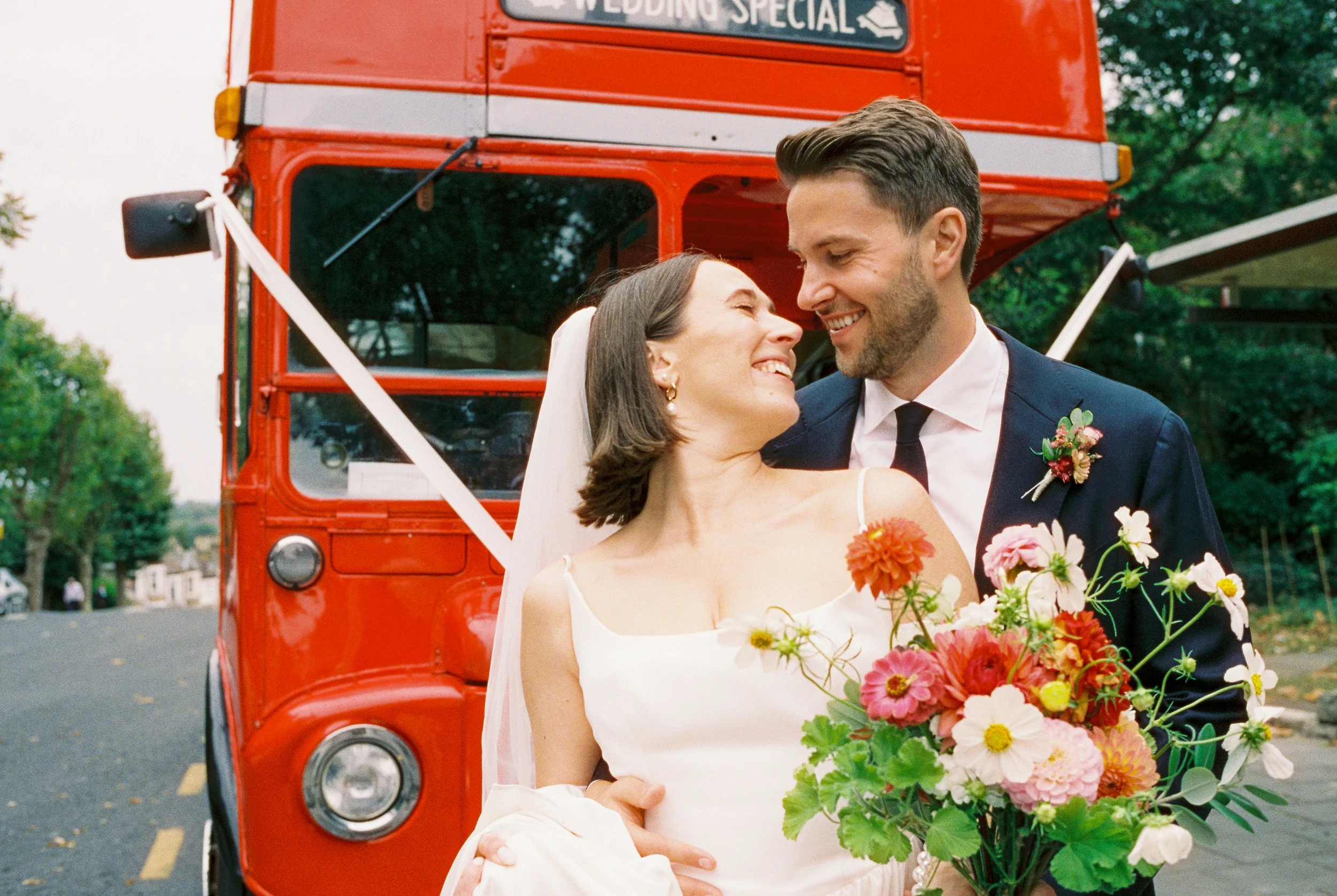 A bride and groom happily embrace in front of a red double-decker bus during their wedding. The bride is holding a bouquet of colorful flowers, and they are smiling at each other.