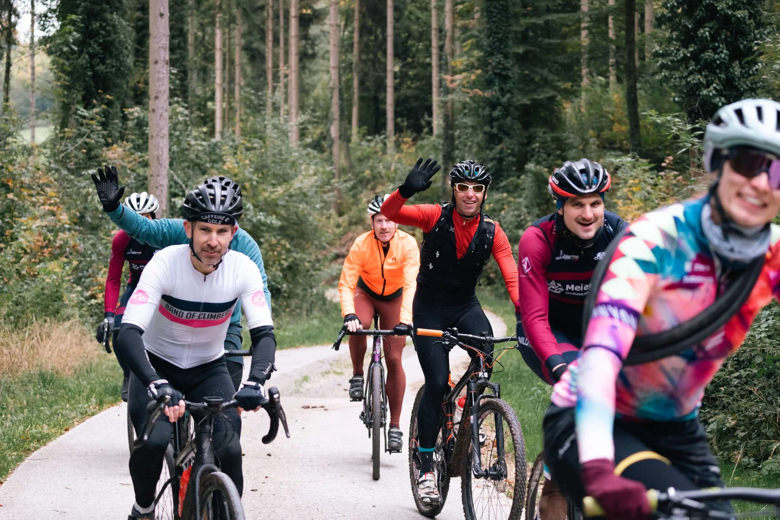 Gruppe von Radfahrern auf einem Waldweg, lächelnd und winkend, umgeben von Bäumen im Hintergrund.