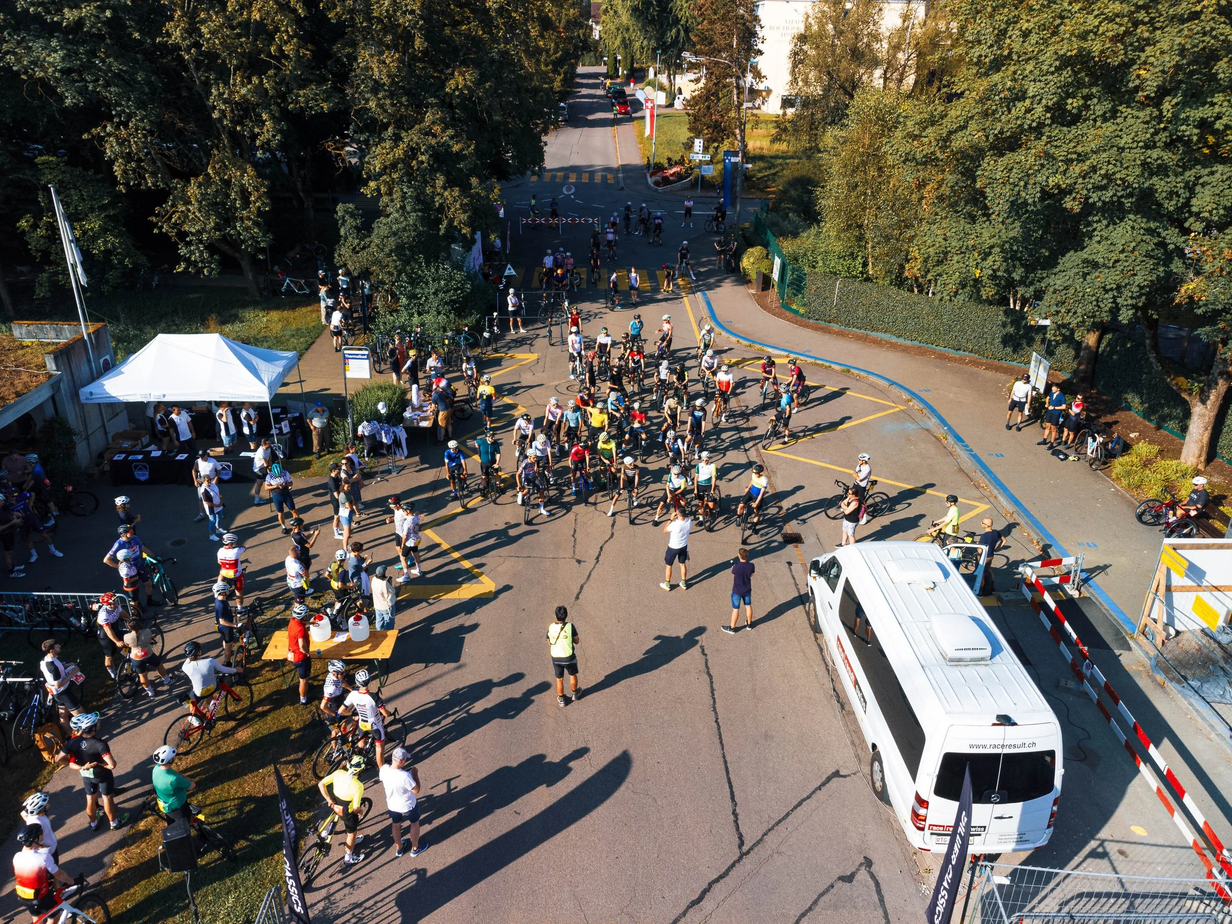 Menschen versammeln sich mit Fahrrädern auf einer Straße, umgeben von Bäumen und einem weißen Zelt. Ein weißer Transporter ist ebenfalls zu sehen.