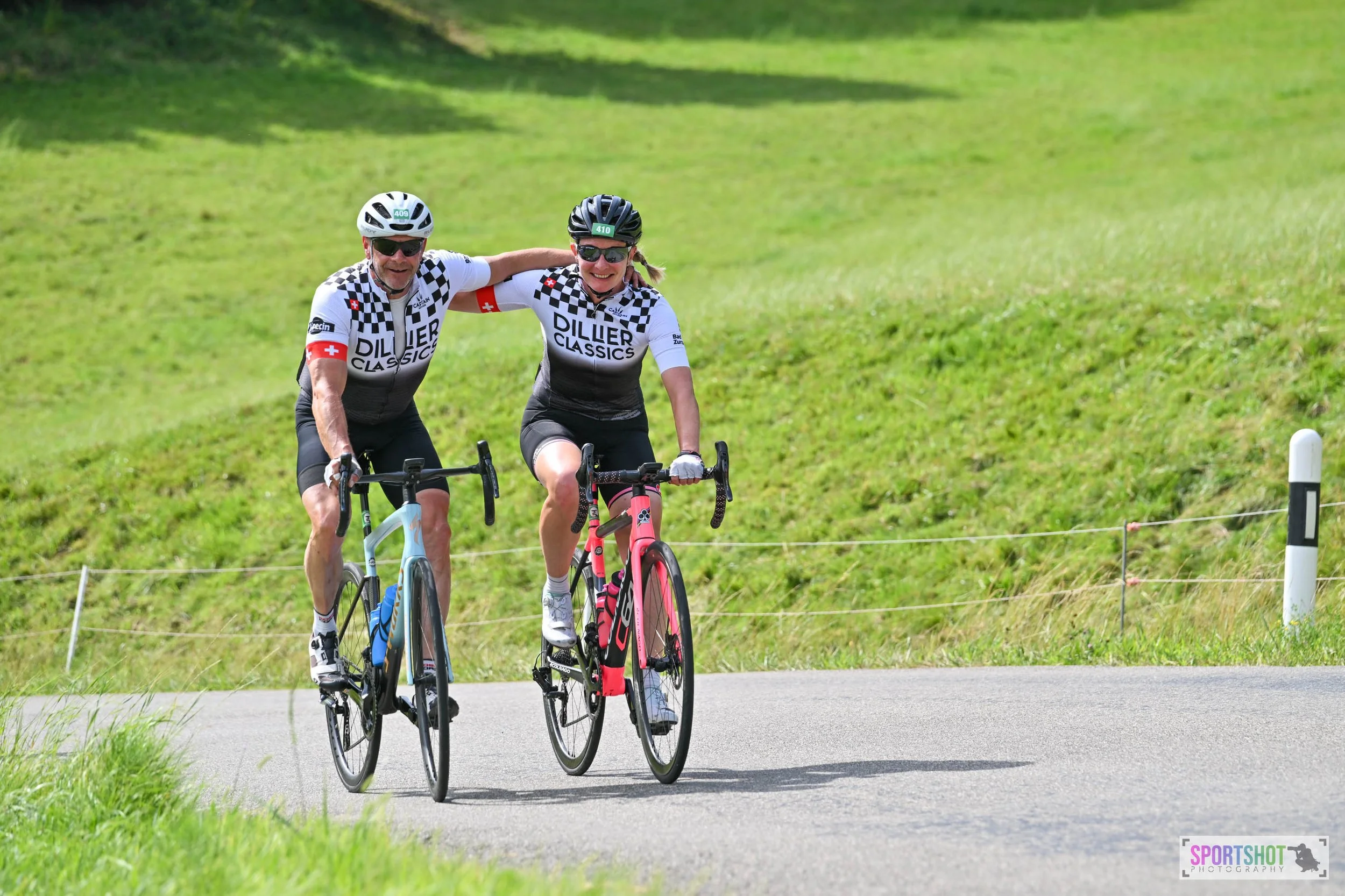 Zwei Radfahrer in der Natur tragen ähnliche Sportkleidung mit dem Schriftzug 'Dillier Classics' und fahren auf einer Straße, einer von ihnen legt den Arm um den anderen. Im Hintergrund ist grüne Landschaft zu sehen.
