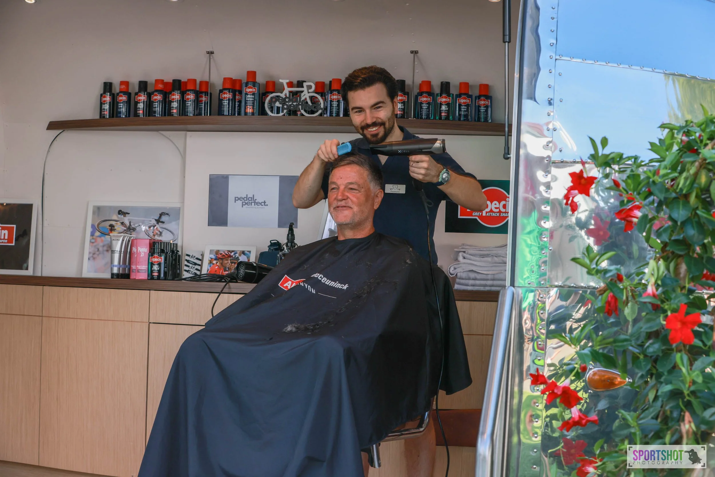 Ein Friseur schneidet einem Mann im Friseursalon die Haare. Im Hintergrund sind Regale mit Haarpflegeprodukten sichtbar. Der Mann trägt eine Schürze mit Markenaufdruck. Rechts im Bild sind Blumen zu sehen.