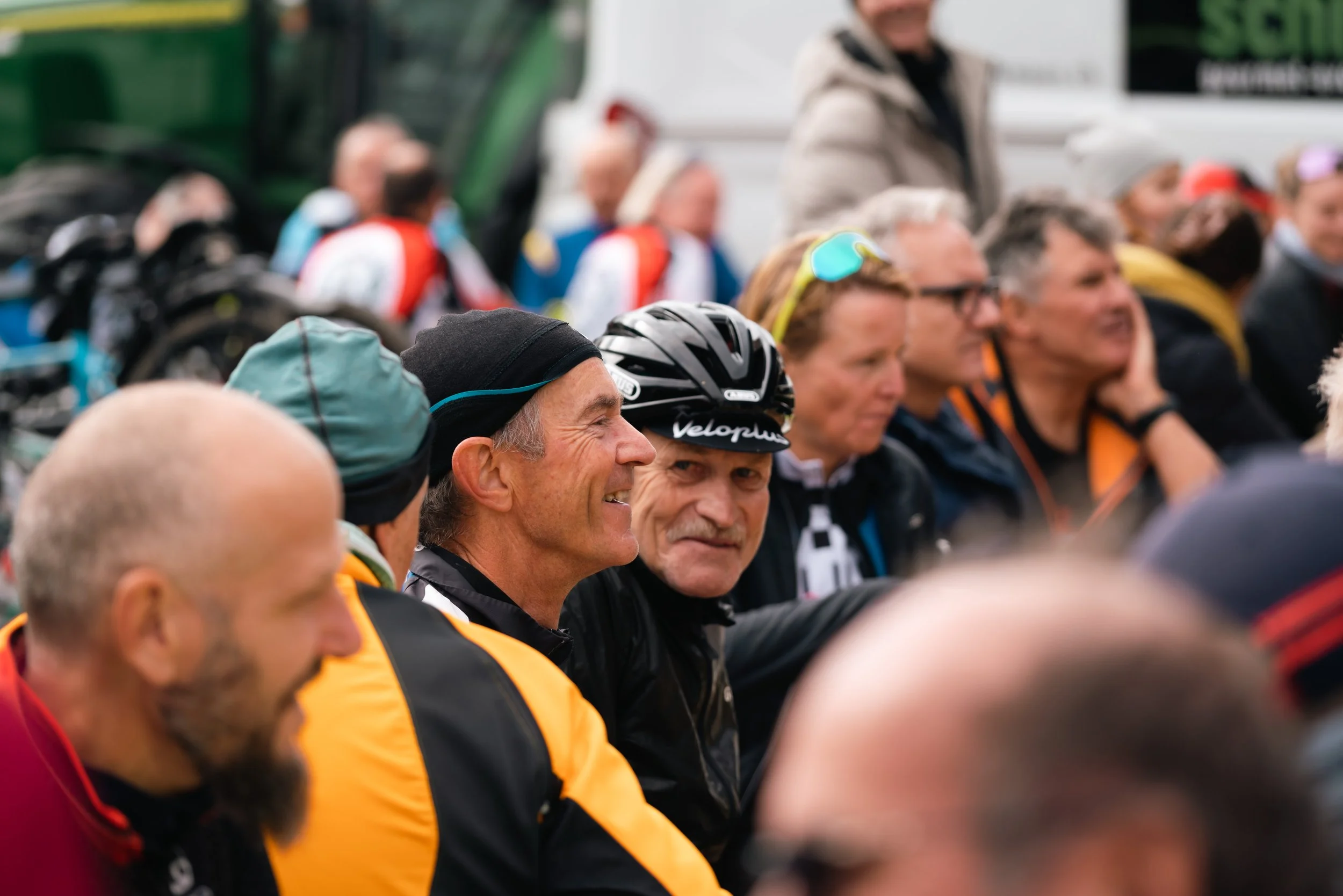 Eine Gruppe von Radfahrern in bunter Kleidung und Helmen, die zusammenstehen und möglicherweise auf ein Radsport-Event warten. Einige Personen lachen und unterhalten sich.