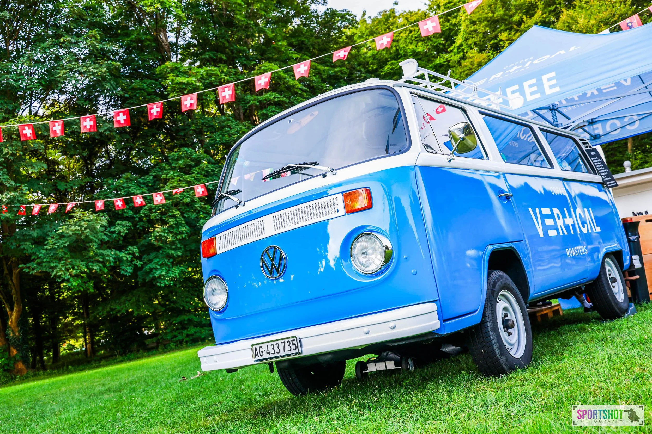 Blauer VW-Bus mit "Vertical Roasters"-Aufschrift auf grünem Gras, umgeben von roten Flaggen mit weißem Kreuz, im Freien vor Bäumen.