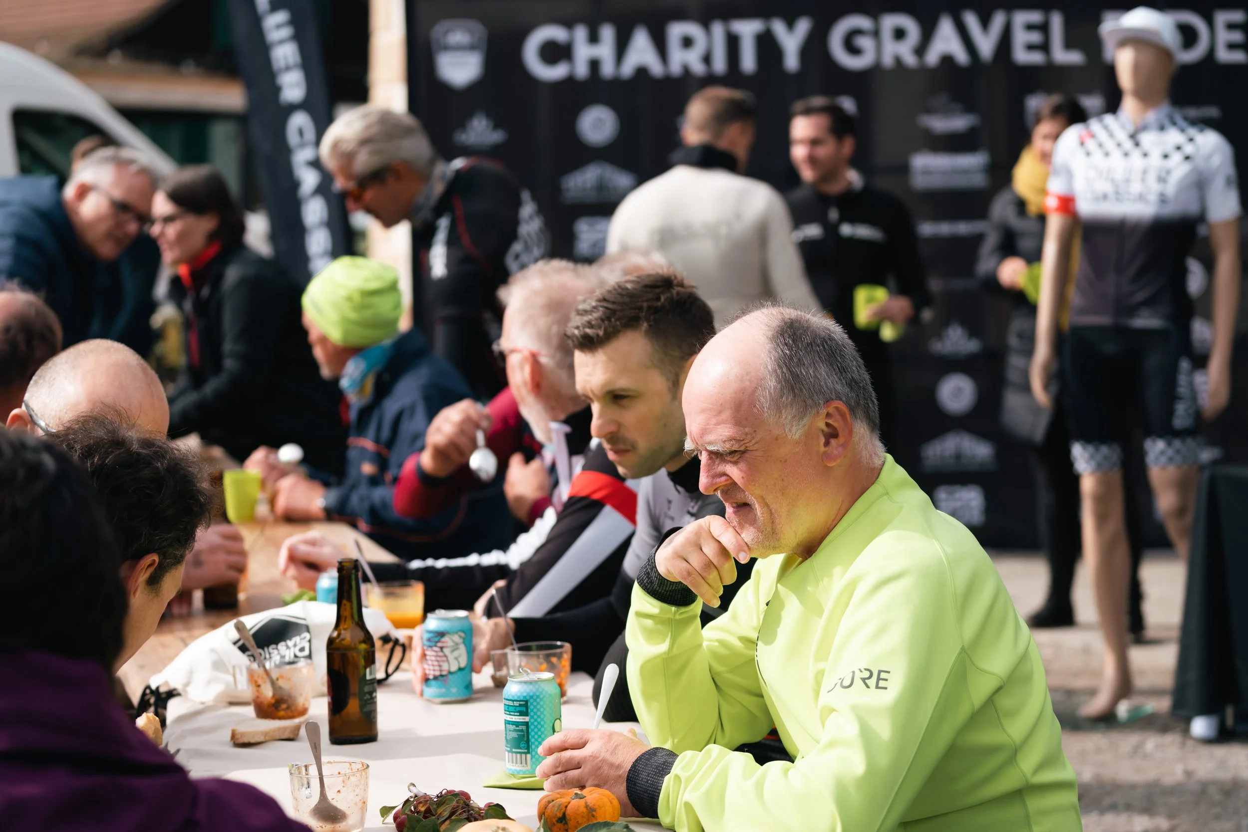 Menschen bei einer Charity-Gravel-Ride-Veranstaltung, die an einem Tisch im Freien sitzen und essen, mit einem Banner und einer Schaufensterpuppe im Hintergrund.