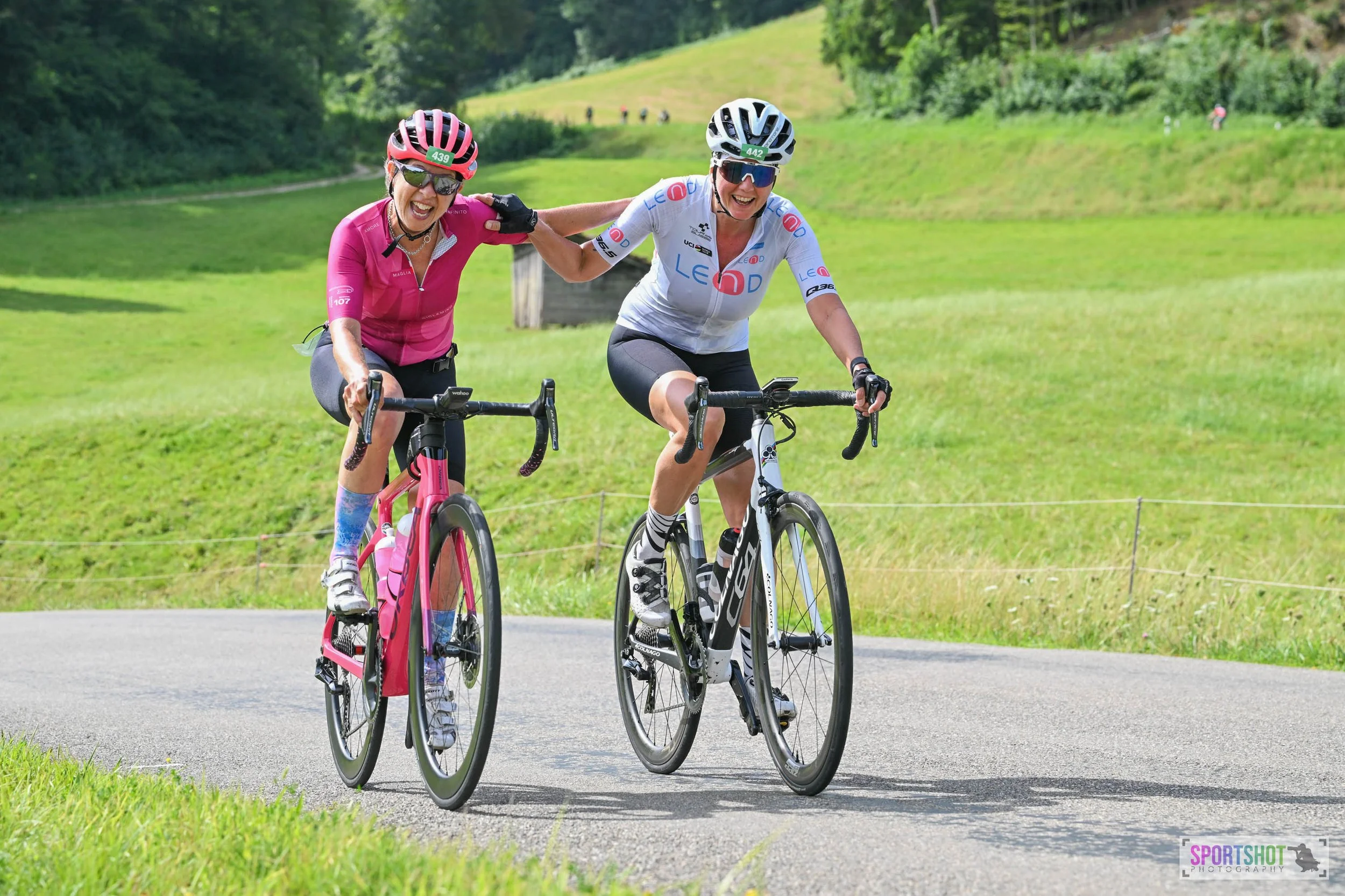 Zwei Radfahrerinnen auf einer Straße in grüner Landschaft. Eine trägt ein pinkes Trikot, die andere ein weißes. Sie lachen und berühren sich freundschaftlich. Im Hintergrund sind Wiesen und Bäume zu sehen.