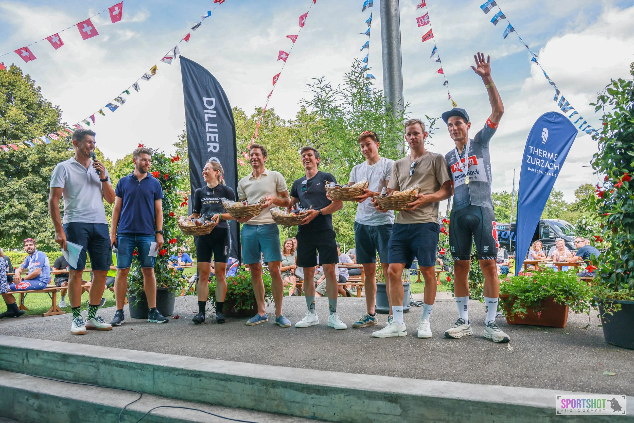 Gruppe von sieben lächelnden Männern und Frauen auf einer Bühne, die Geschenkkörbe halten. Im Hintergrund sind Dekorationen mit Schweizer Flaggen zu sehen. Rechts im Bild stehen zwei Banner mit den Aufschriften „Dillier“ und „Therme Zurzach“. Im Vord