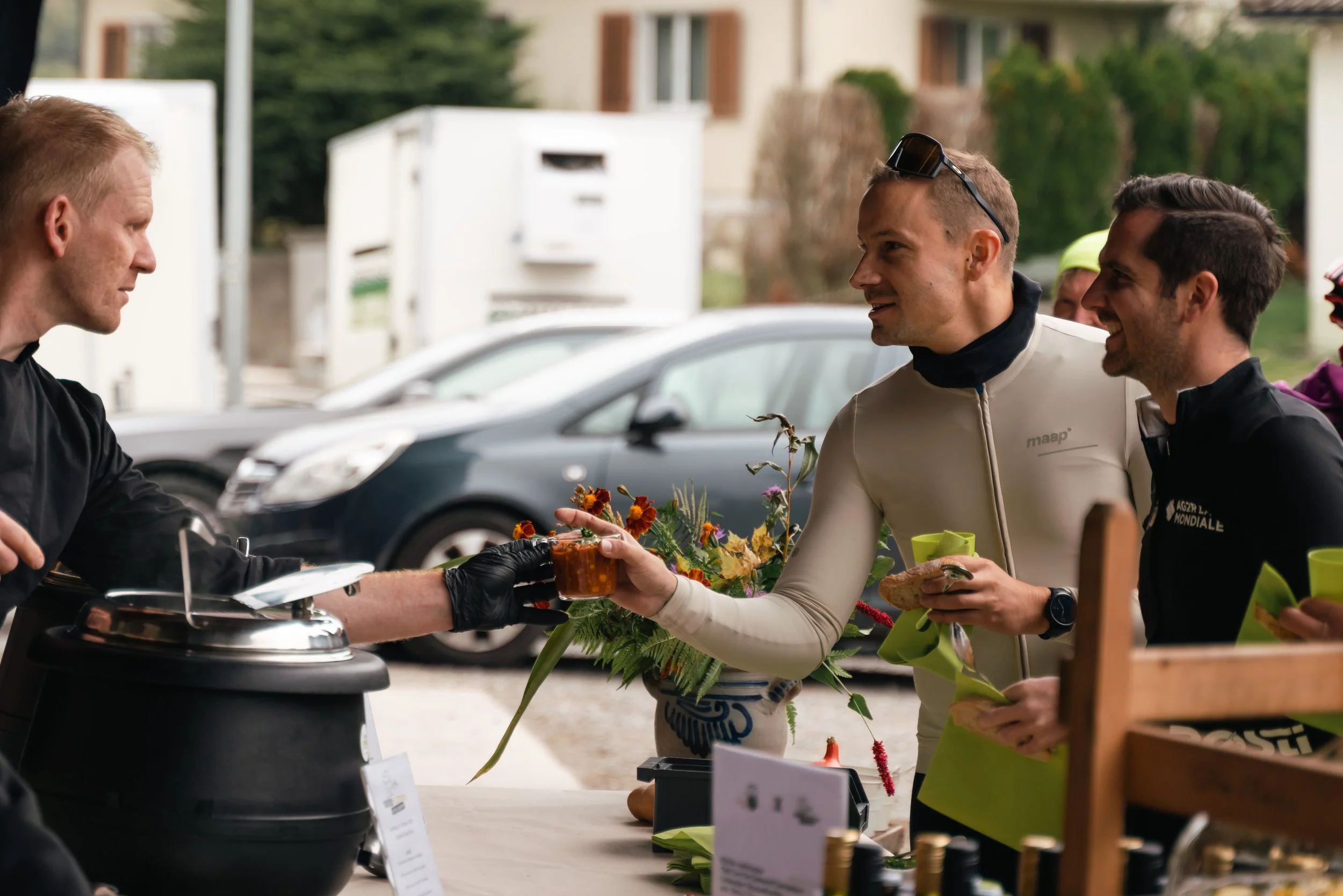 Drei Männer an einem Essensstand, einer serviert eine Suppe, die anderen lächeln und halten Becher und Essen. Im Hintergrund sind Autos und Häuser sichtbar.