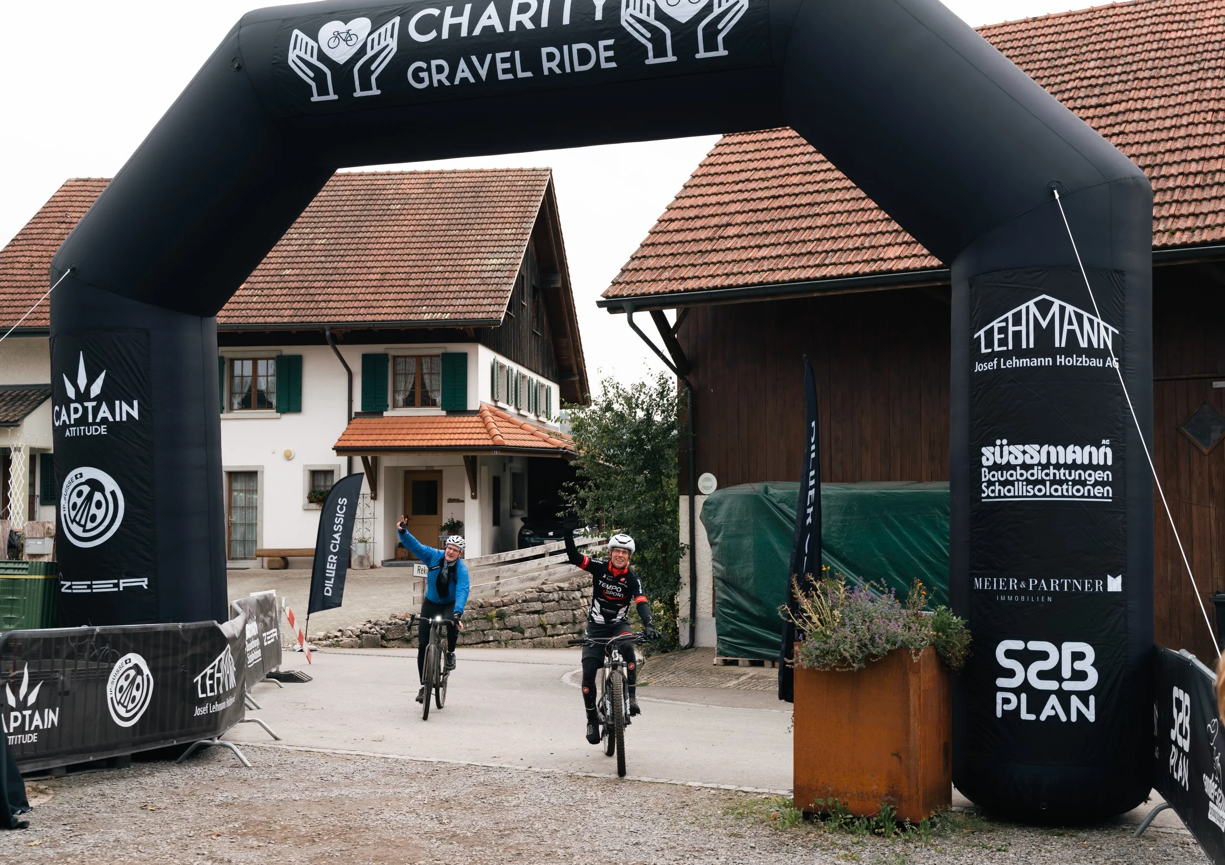 Die Radfahrer heben ihre Arme siegreich in die Luft und fahren unter dem Zielbogen des Charity Gravel Rides hindurch. Sie freuen sich nun auf ein wohlverdientes Finisher Meal vom Catering Sonderschmaus.