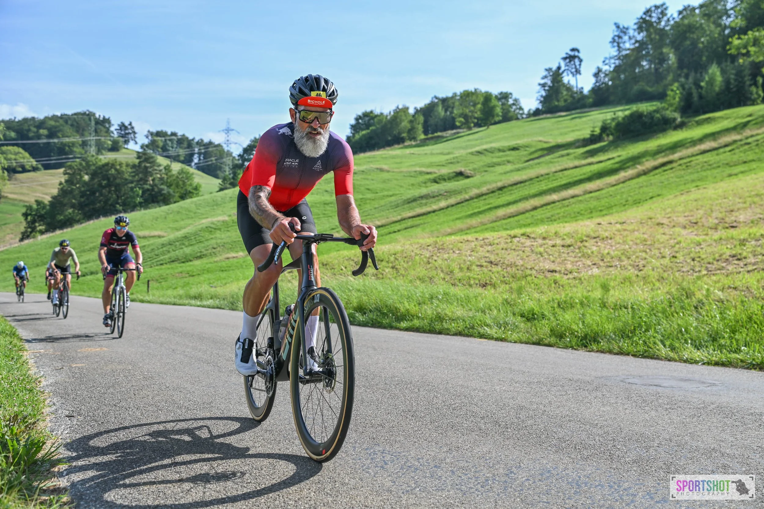 Eine Gruppe von Radfahrern fährt auf einer Landstraße bergauf. Im Vordergrund ist ein Mann mit roten und schwarzen Trikot zu sehen, der ein modernes Rennrad fährt. Die Landschaft ist grün und hügelig, mit Bäumen im Hintergrund unter klarem, blauem Hi