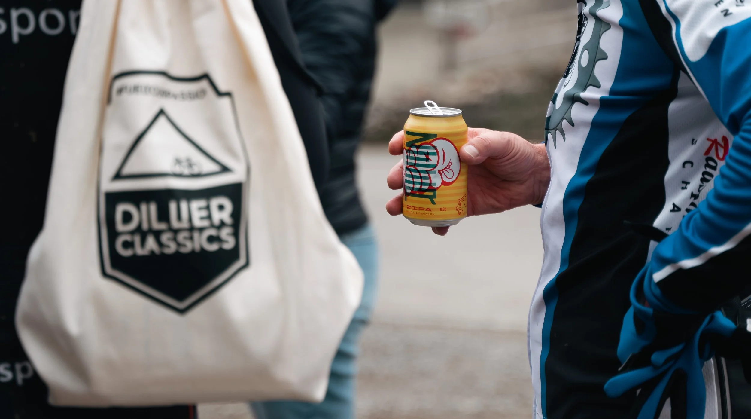 Person hält Getränkedose mit bunter Gestaltung, trägt ein Trikot und hat eine Tasche mit der Aufschrift 'Dillier Classics'. Hintergrund unscharf.